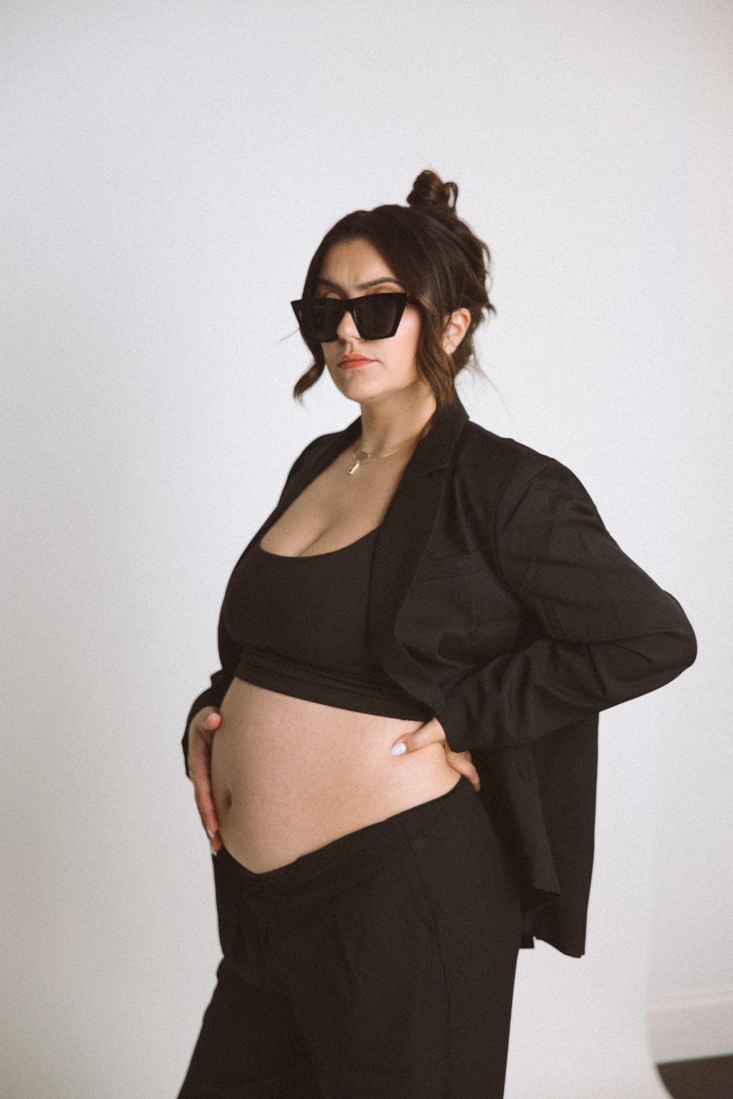 Modern maternity portrait with woman in black blazer and sunglasses against white background.