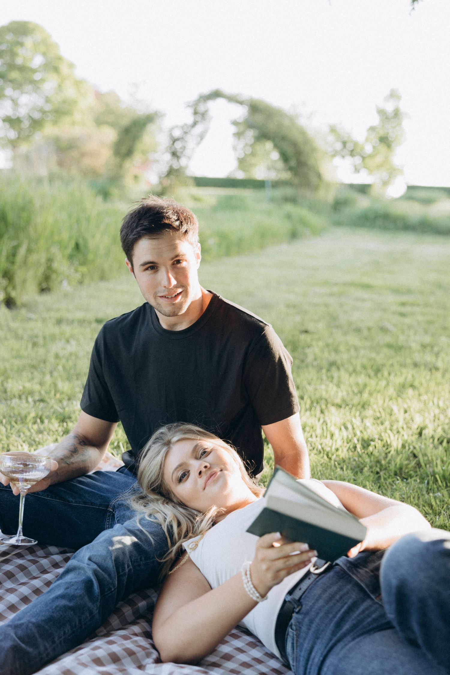 A couple relaxes on a picnic blanket in a sunny field with drinks and a book.