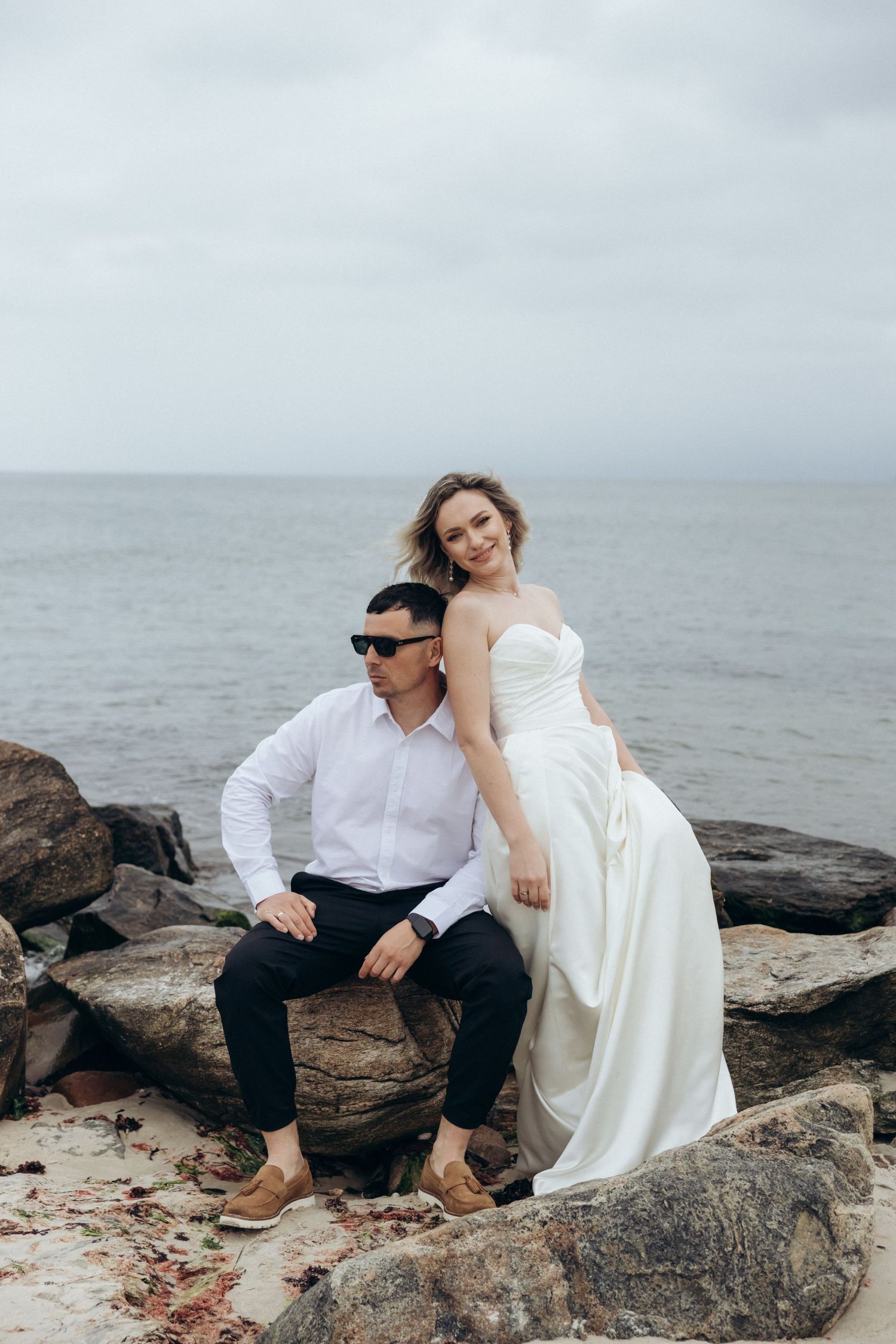Wedding couple sitting together on large coastal rocks by the ocean.