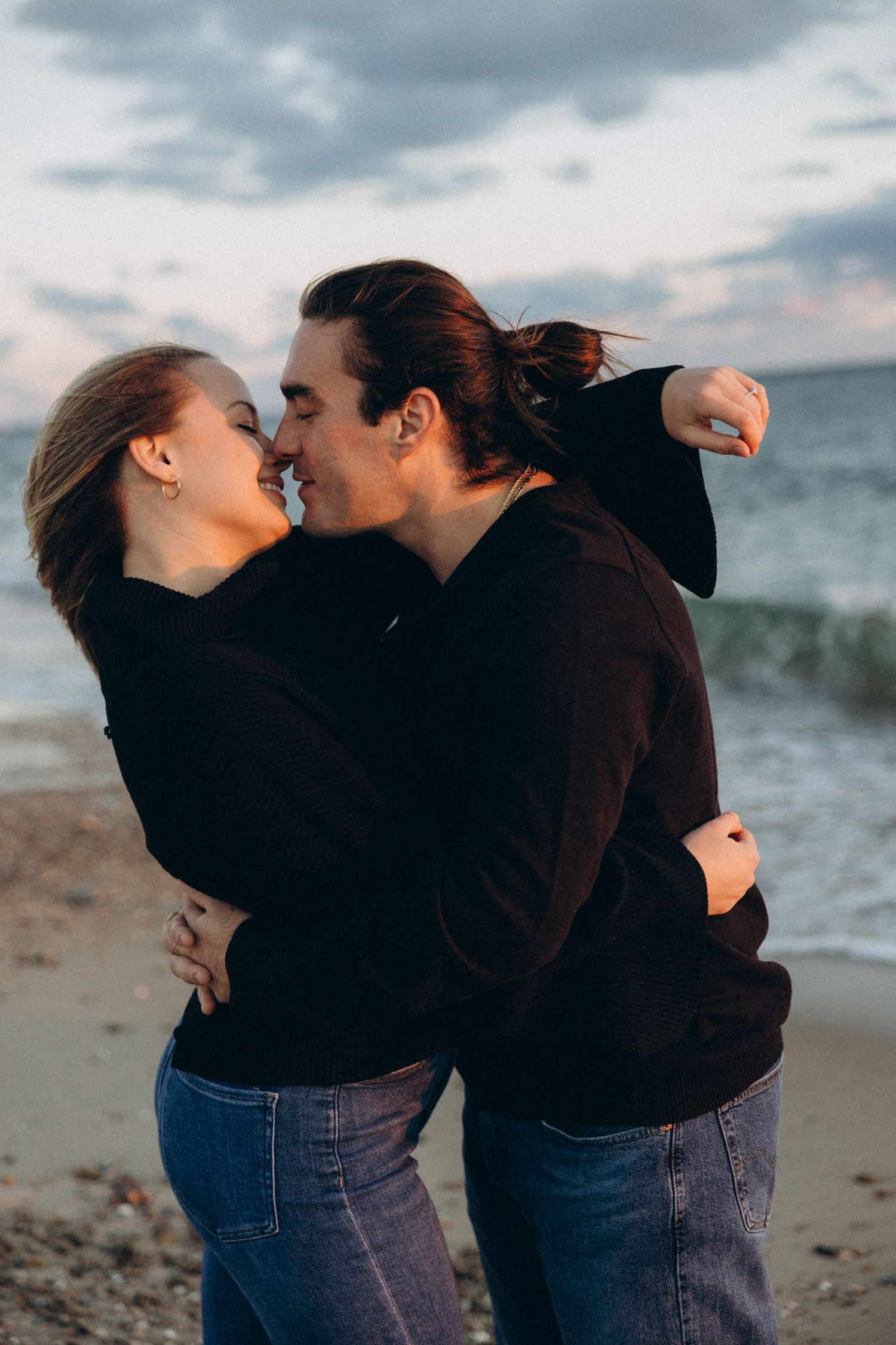 Intimate beach photoshoot showing a couple in black sweaters sharing affectionate moments.
