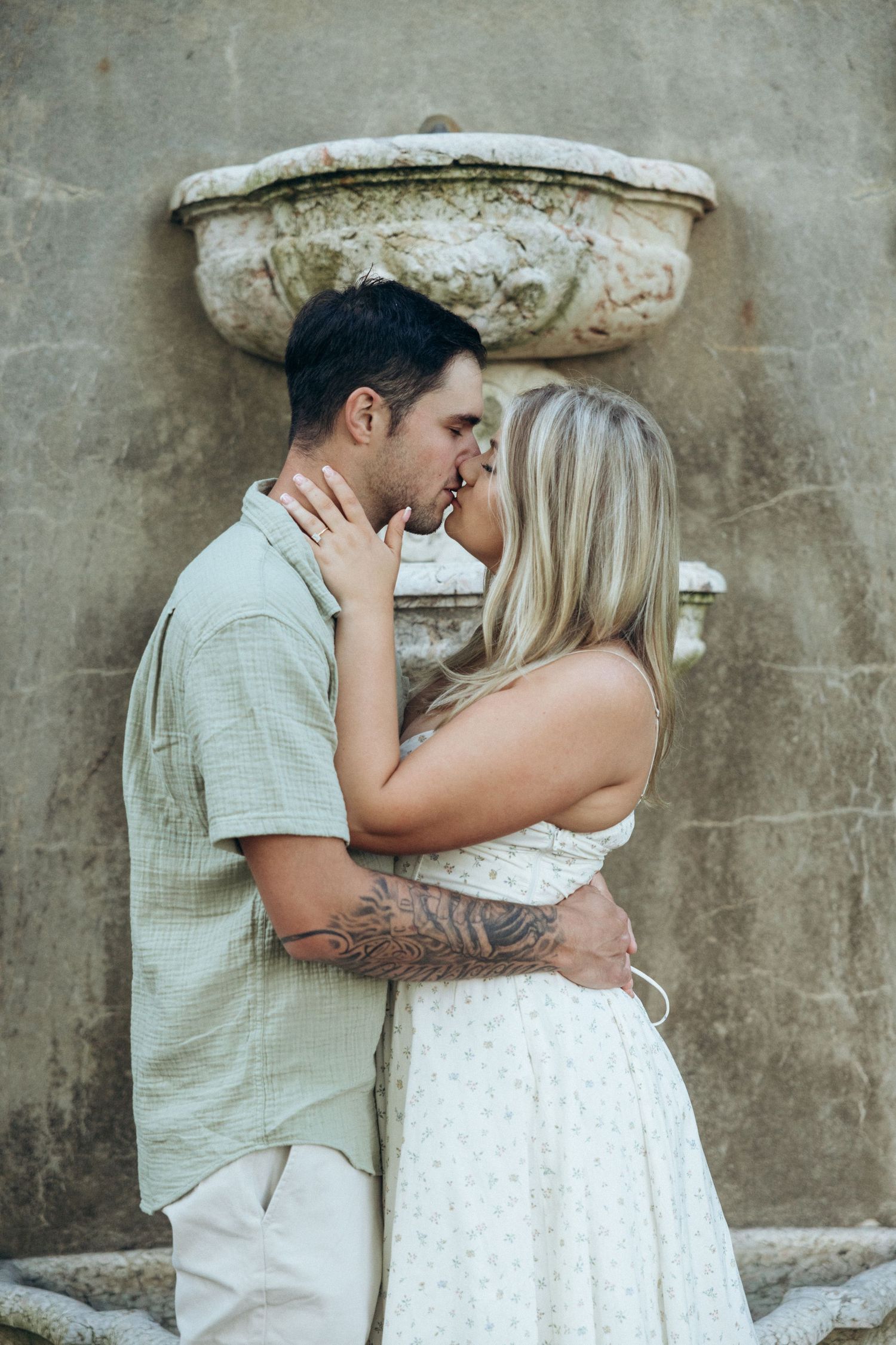 A couple in light summer clothing embracing against a weathered stone wall with decorative details.