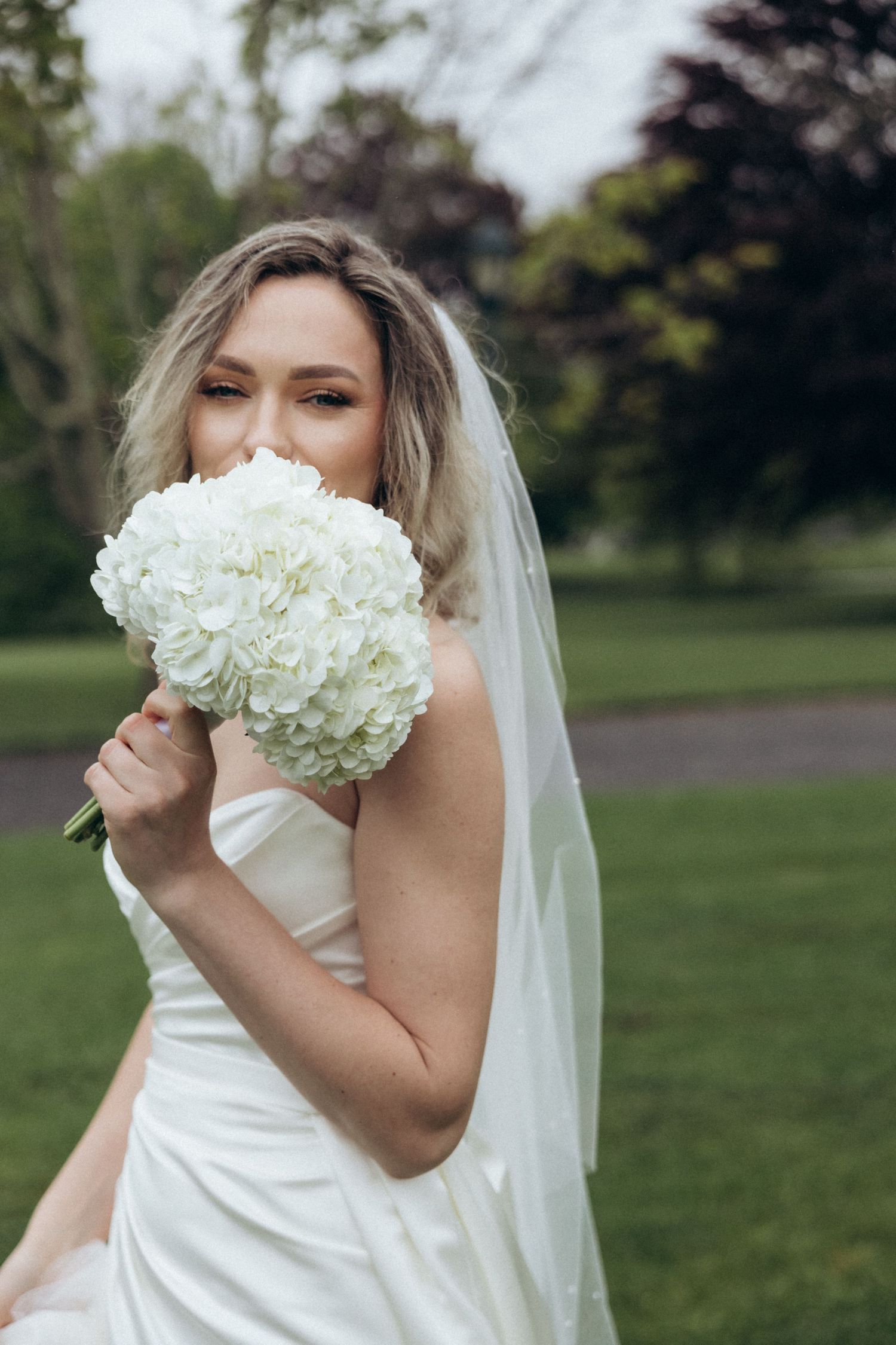 A white hydrangea bouquet held up in front of a bride wearing a white wedding dress.