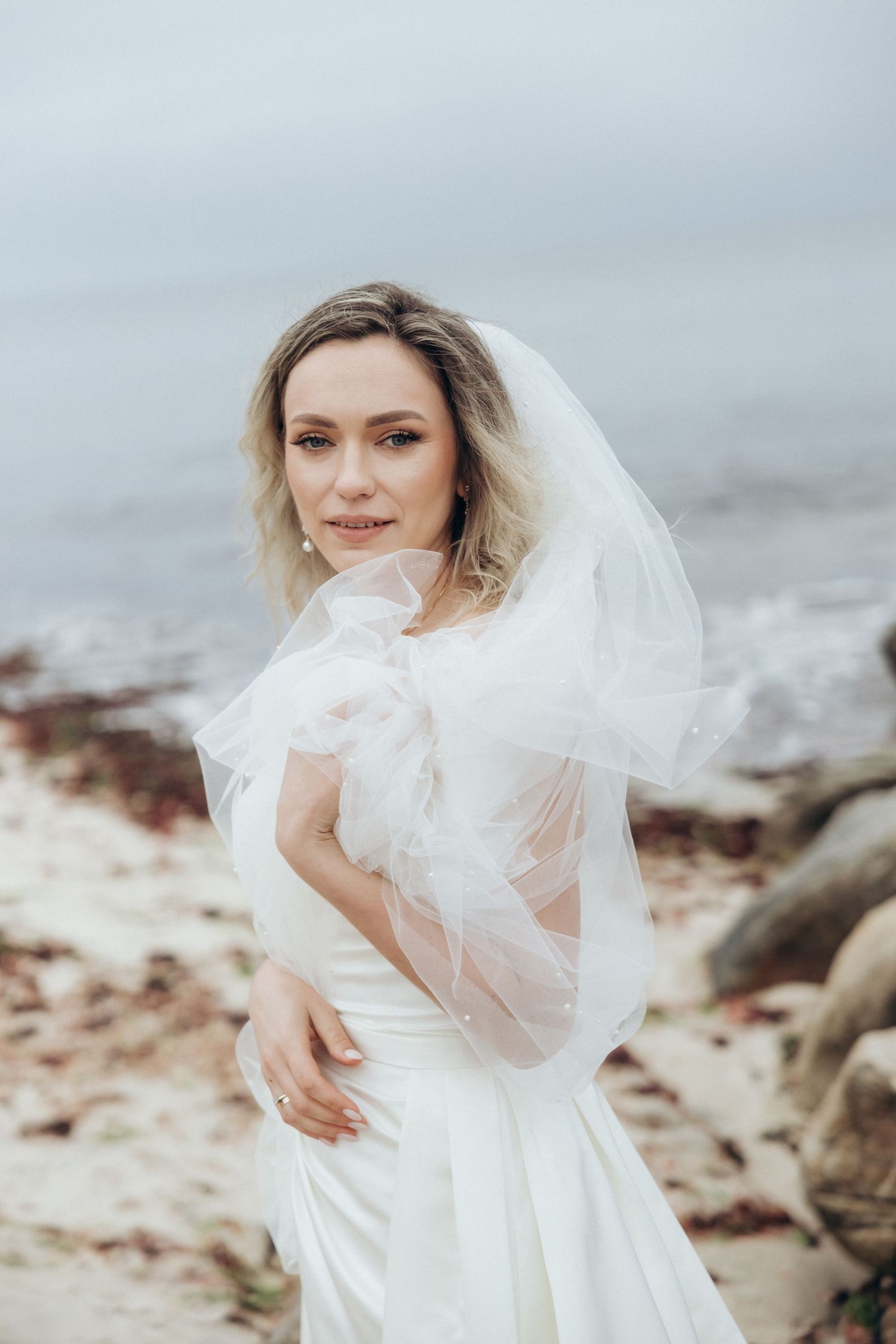 Flowing white wedding veil catches the ocean breeze on a rocky beach.