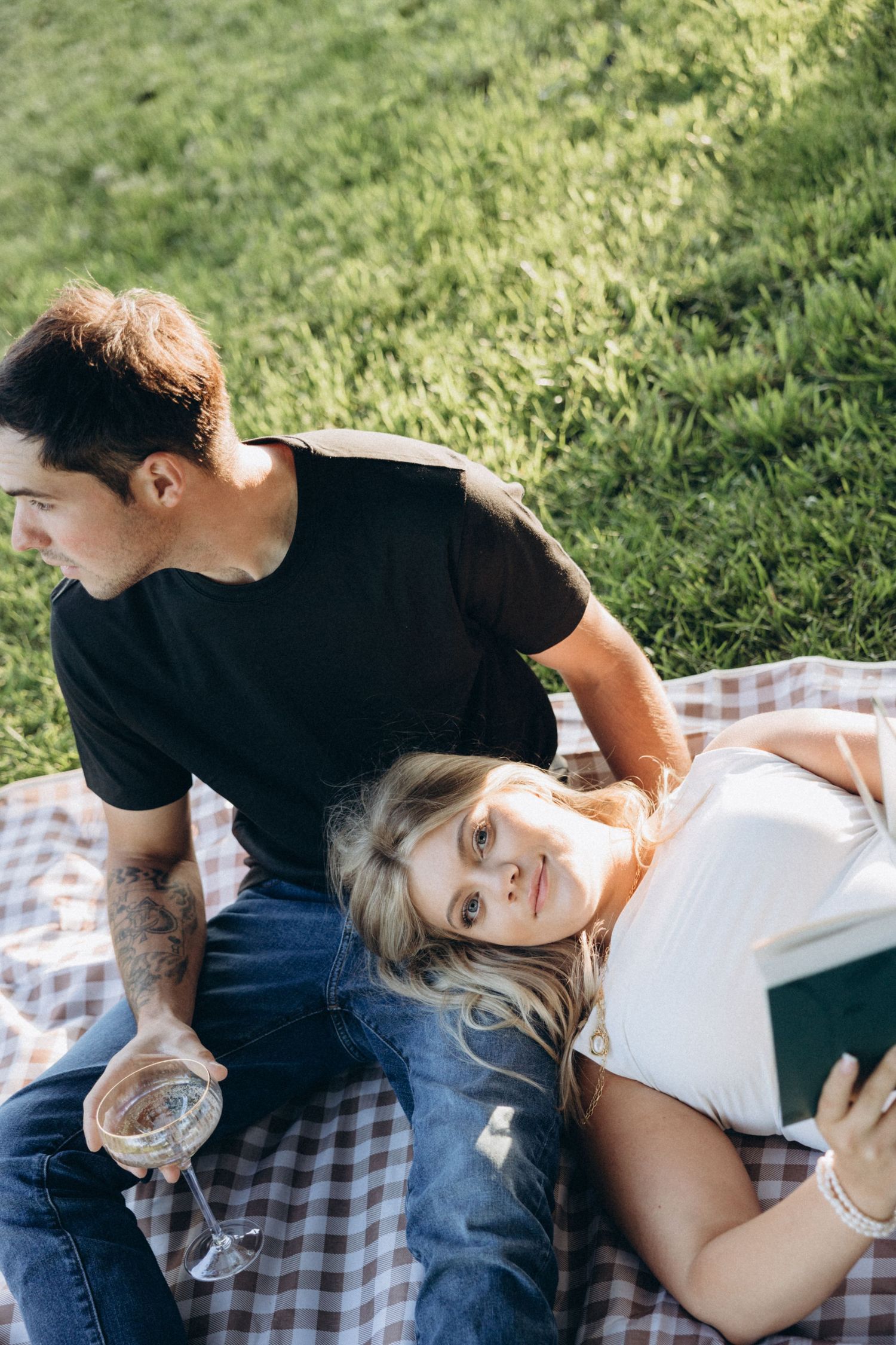 A romantic couple lying together on a checkered picnic blanket in the grass on a sunny day.