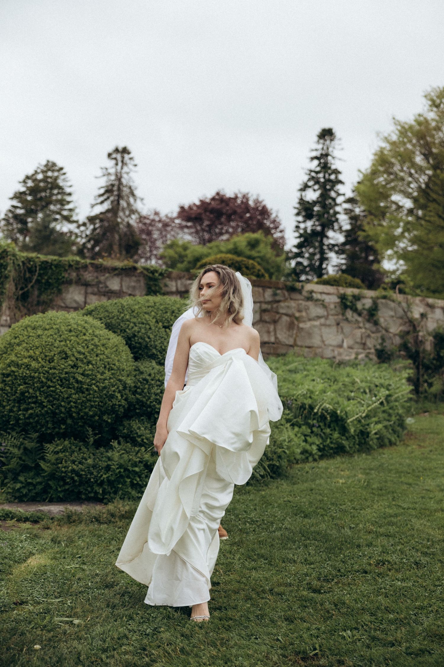 A bride in a flowing white dress moves gracefully through a garden landscape.