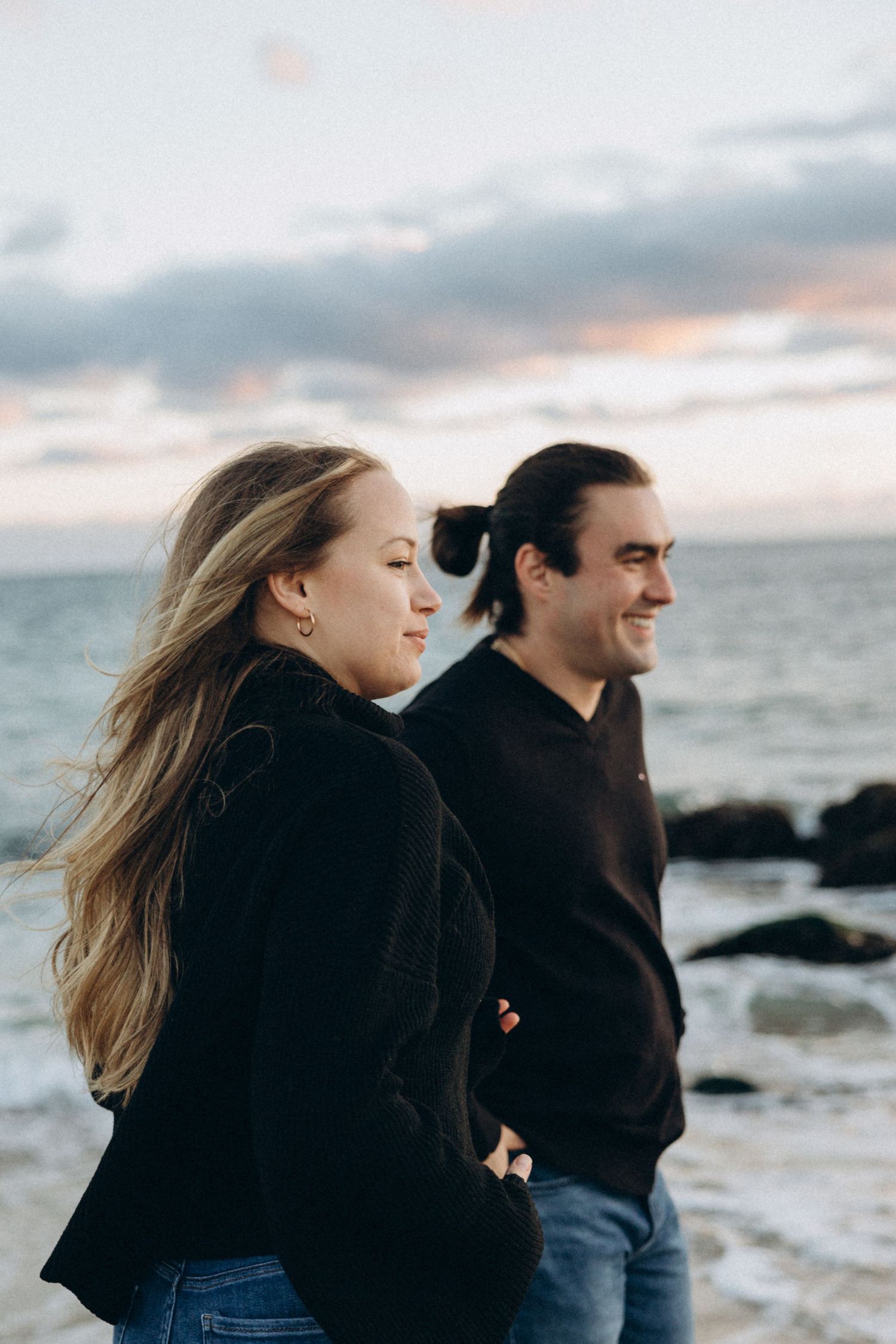 Couple in dark clothing walking together along a rocky beach during golden hour.
