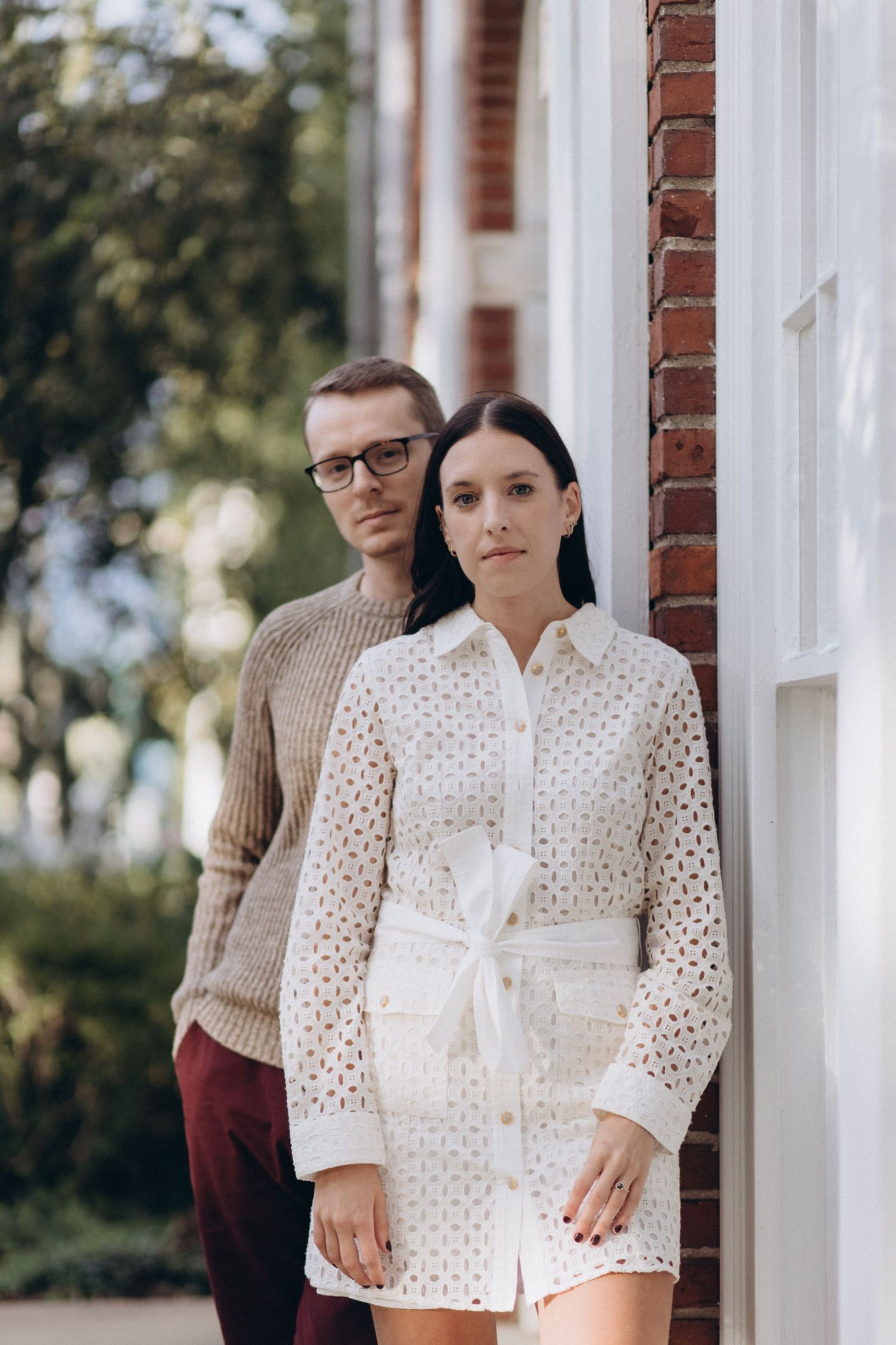 A stylish couple poses against a brick wall wearing coordinated outfits in muted tones.