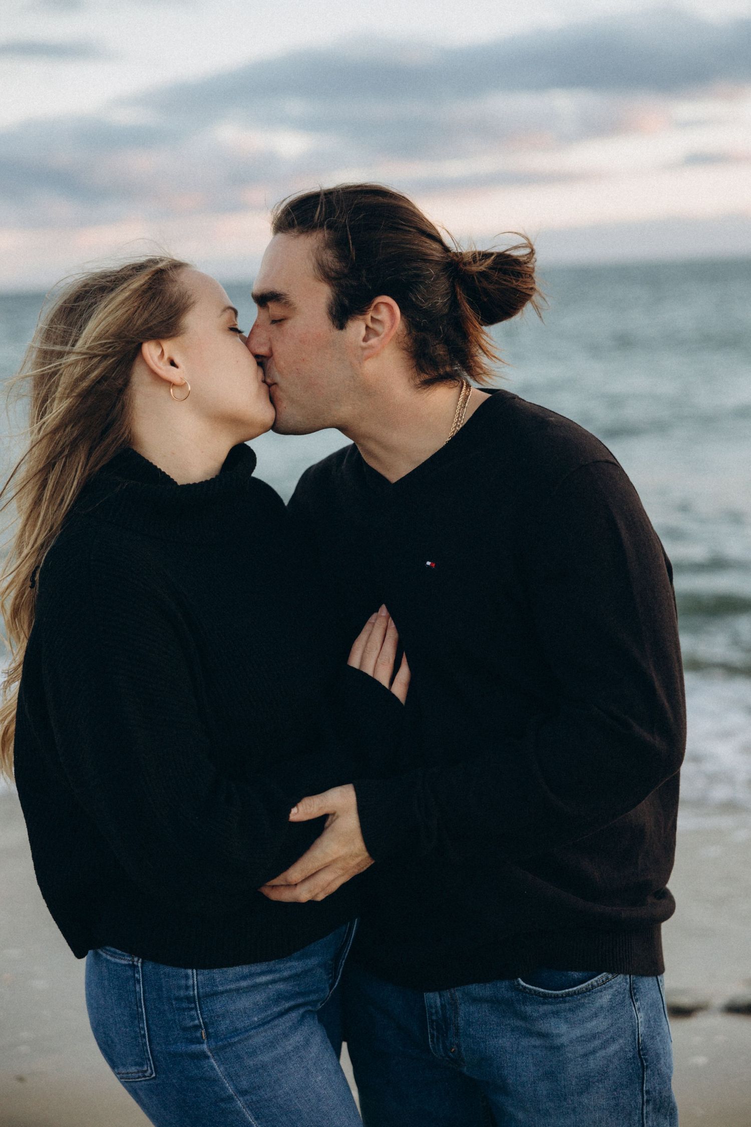 Romantic couple in dark clothing embracing on a beach with waves in the background.