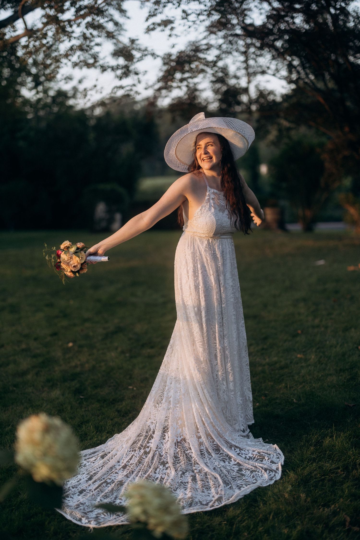 Bride in flowing lace dress and wide-brimmed hat poses with bouquet in golden evening light.