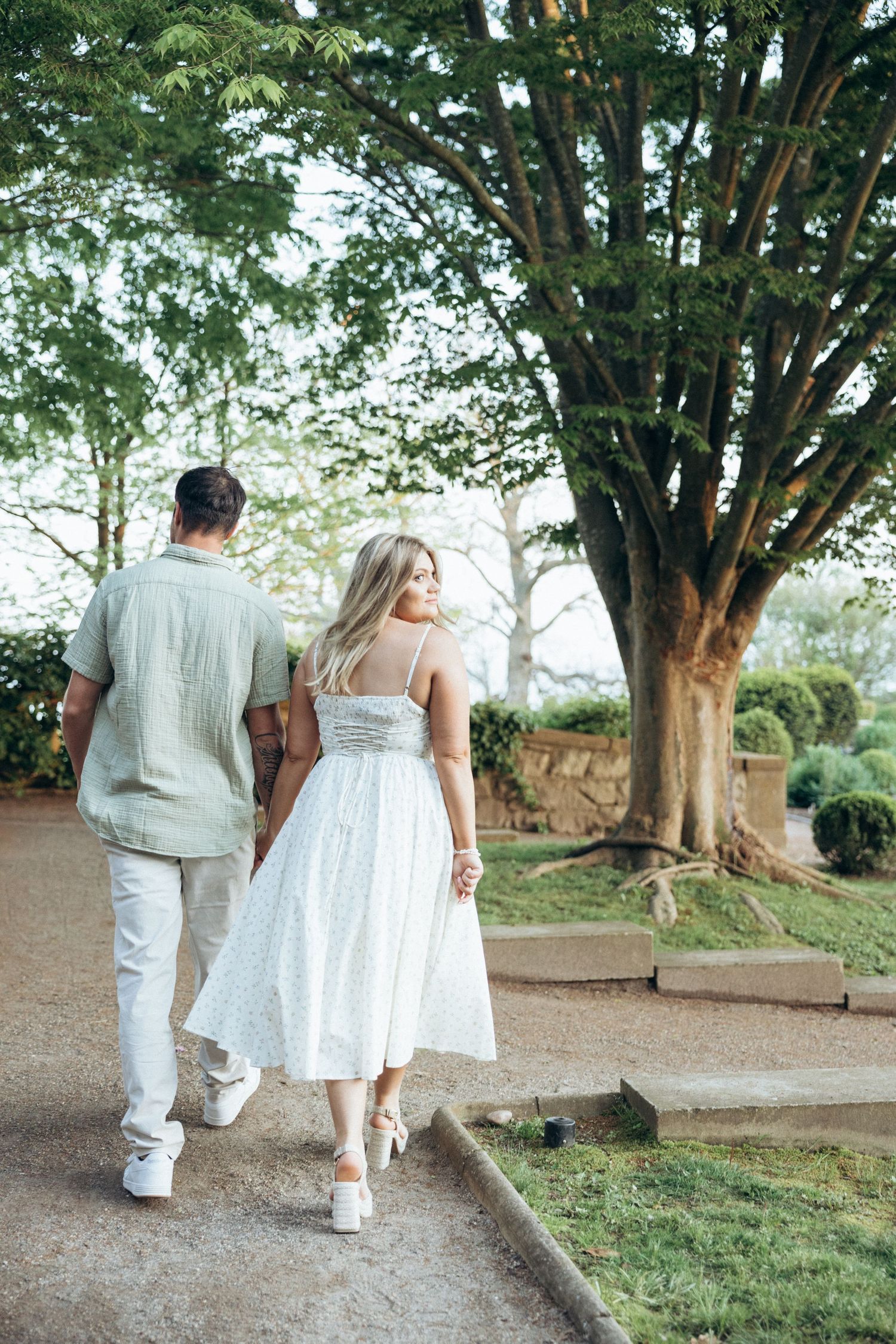 A couple in coordinated white outfits walking along a tree-lined path in a garden.