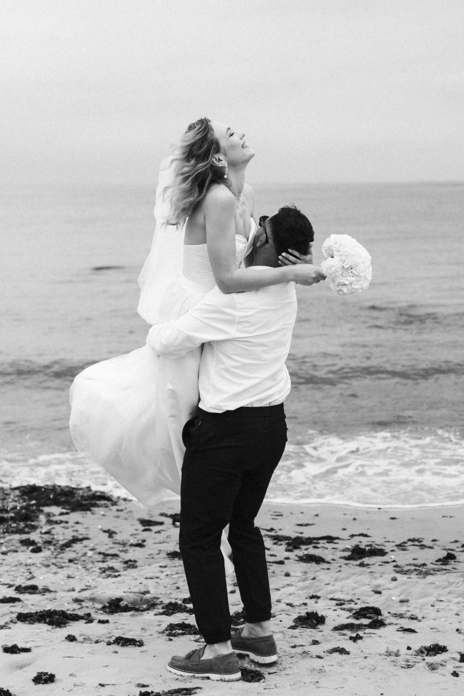 Couple in wedding attire playfully posing on a beach with crashing waves.