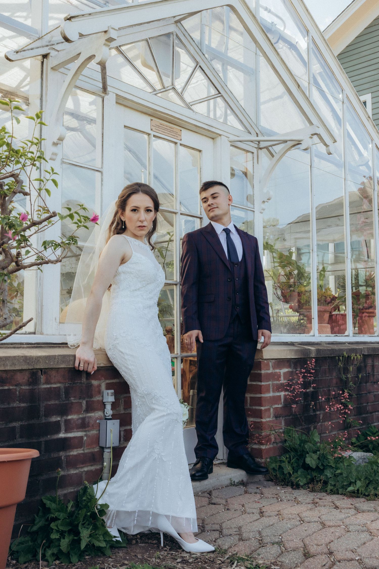 An elegant wedding couple poses in front of a bright greenhouse structure with brick walls.