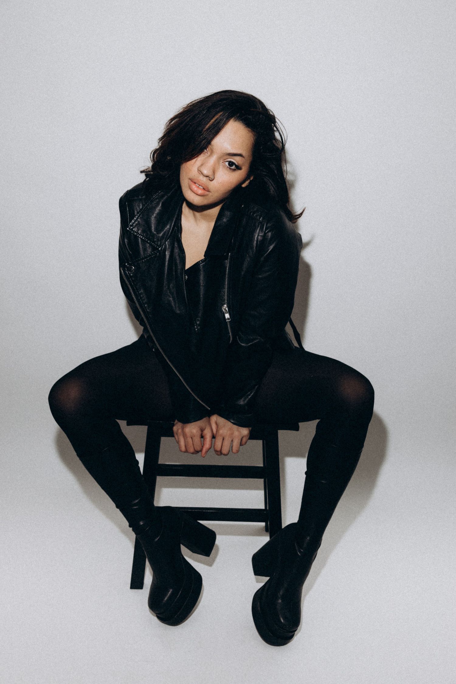 Model in all-black ensemble sits on wooden stool against white wall wearing leather jacket and boots.
