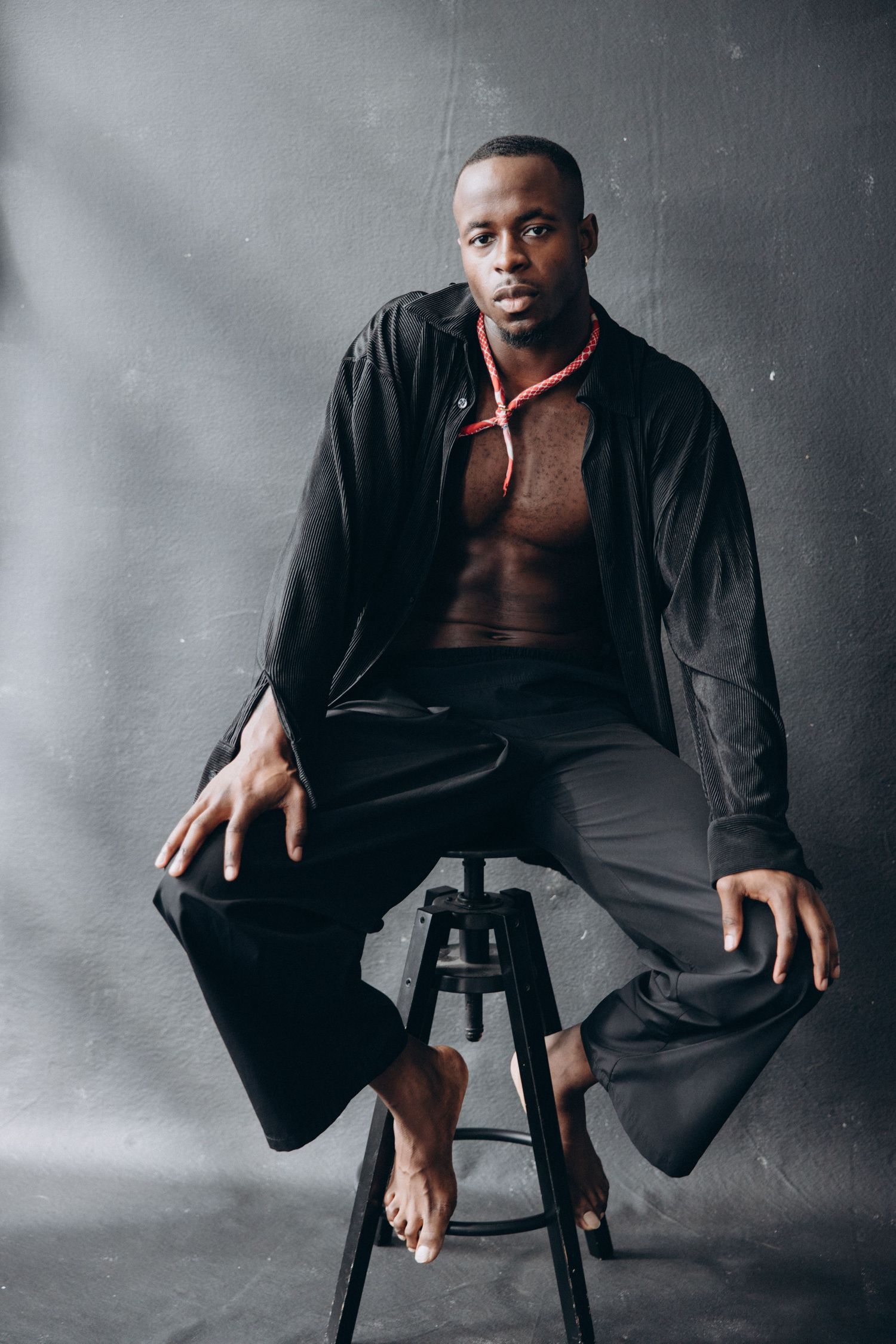 Model in black jacket sits barefoot on stool against gray backdrop in studio setting.