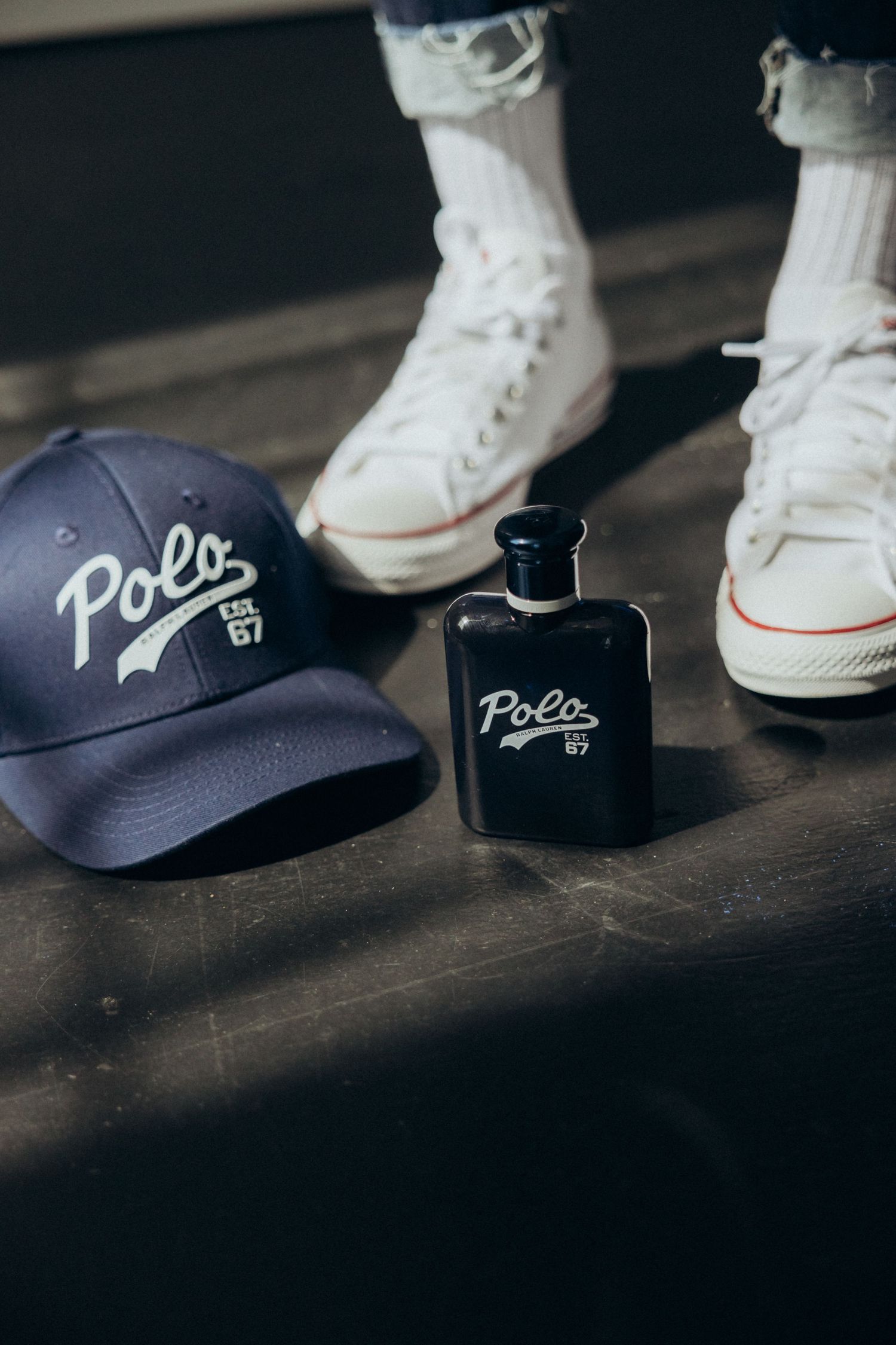 Navy baseball cap, black perfume bottle and white sneakers arranged on dark wooden surface.