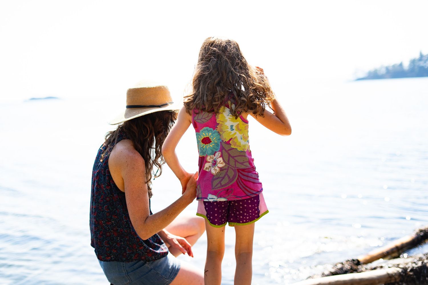 Woman kneeling beside young girl at water's edge, both looking out at calm lake on a sunny day.