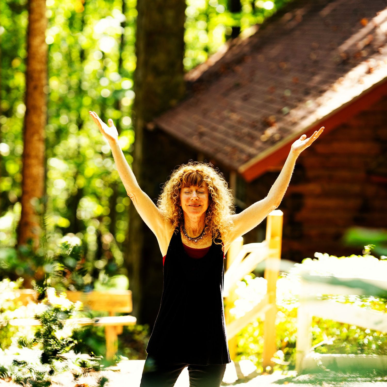 A photo of a woman with orange curly hair wearing dark clothing, her hands are up toward the sky and there is greenery behind her.
