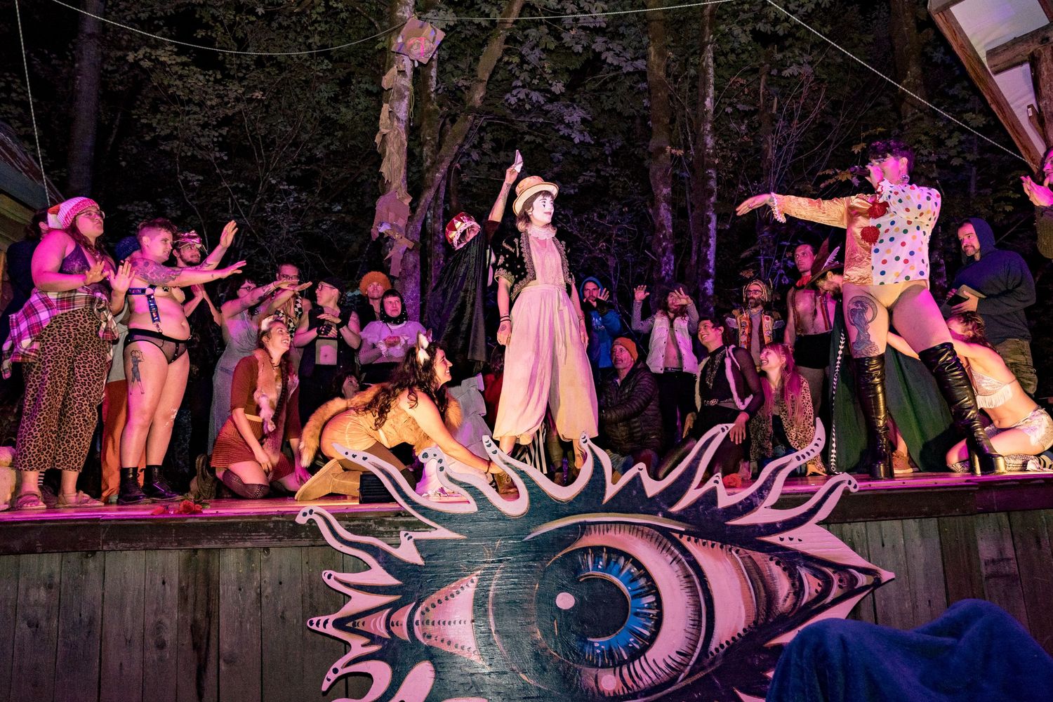Colorful outdoor festival performance with dancers on stage featuring a large eye mural, surrounded by crowd at night.