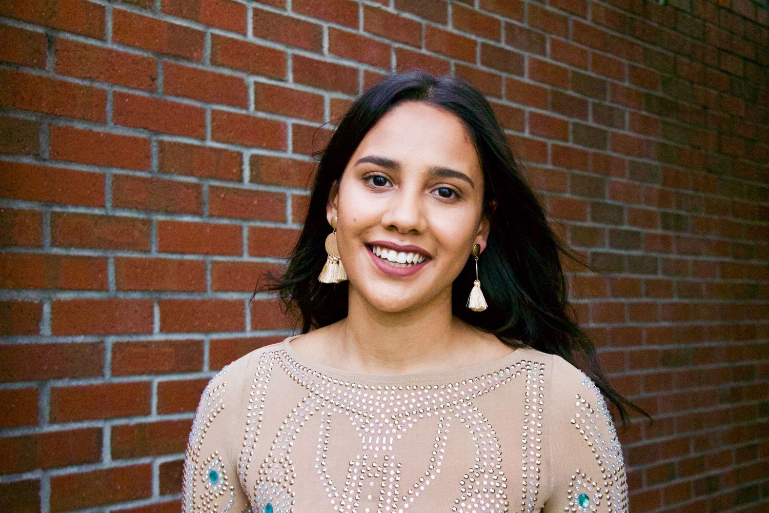 Smiling young woman with dark hair wearing gold earrings and beige embroidered top, posing against a red brick wall.