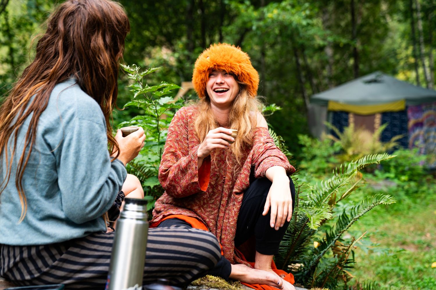 Two women laughing and enjoying drinks outdoors in a lush forest campsite setting.
