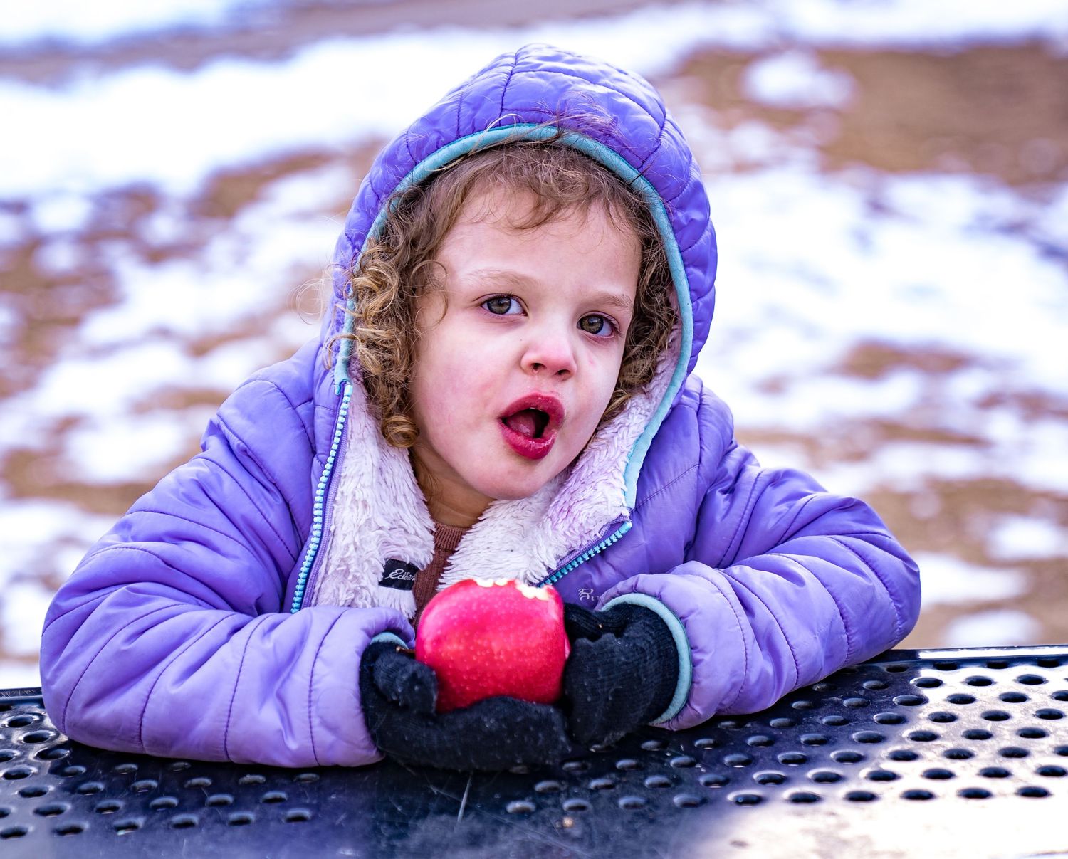 A person in a purple winter coat lies playfully in the snow while making silly expressions.