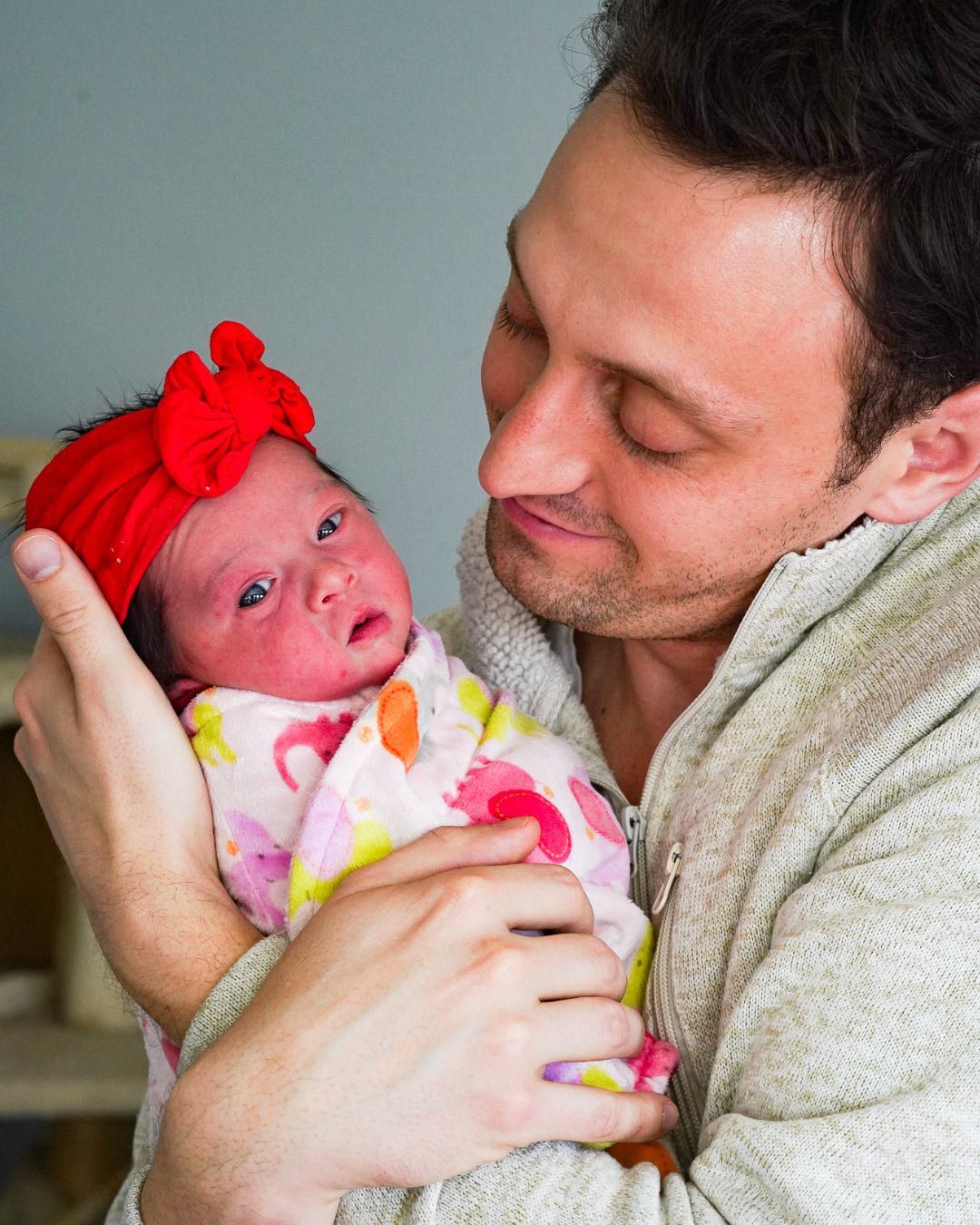 A man tenderly holds a newborn baby girl wearing a red bow headband, wrapped in a colorful floral blanket.