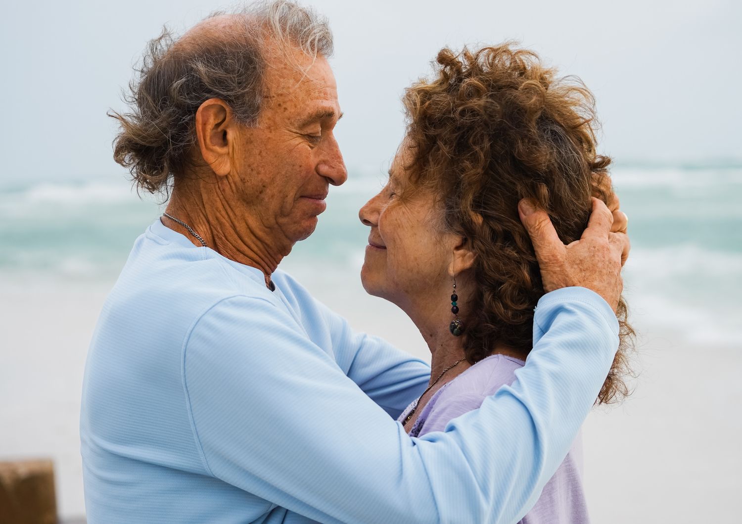 An elderly couple shares a romantic embrace on a beach, gazing into each other's eyes on a cloudy day.