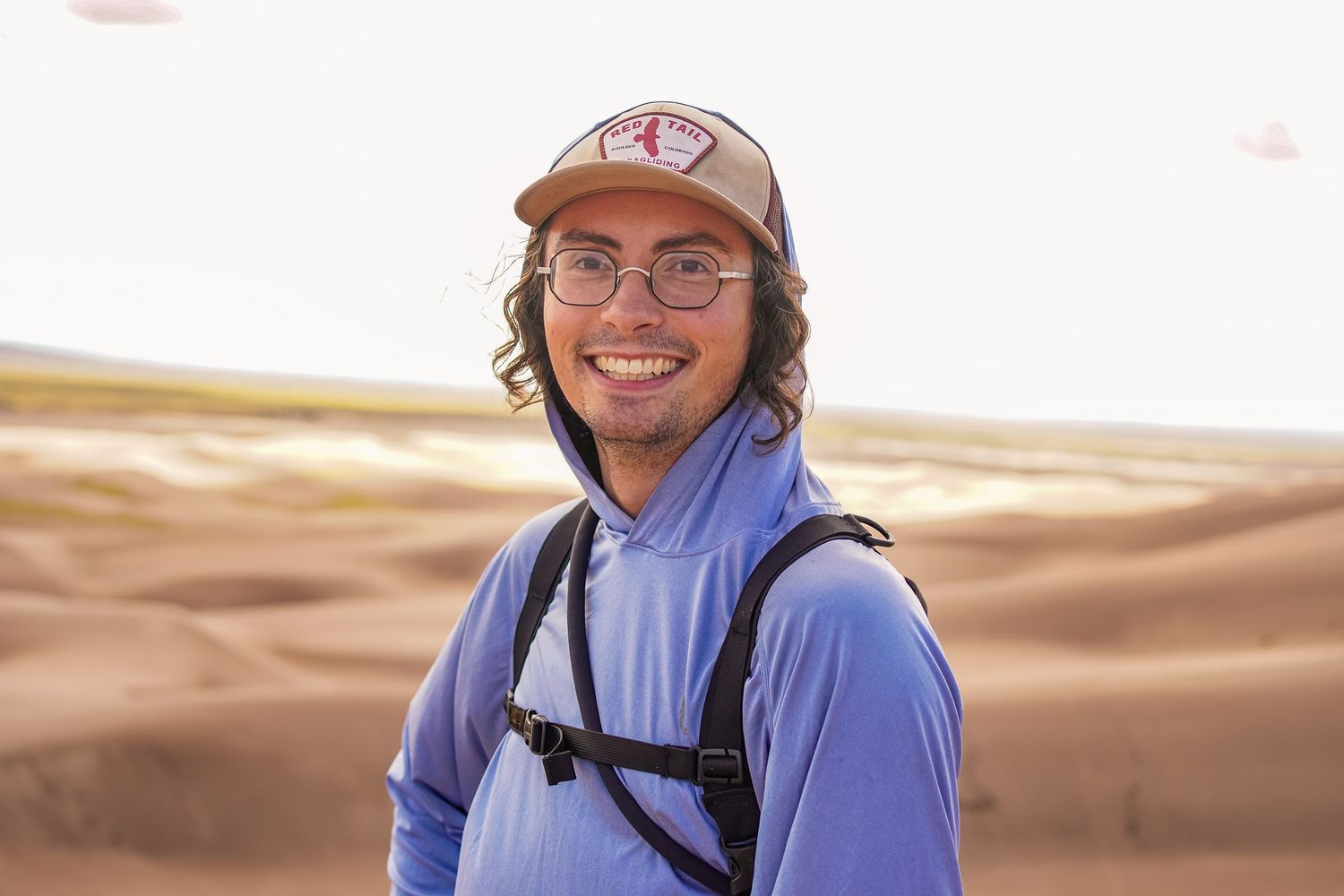 A photo of a guy wearing a hat with a blue sweater, he has curly brown hair.