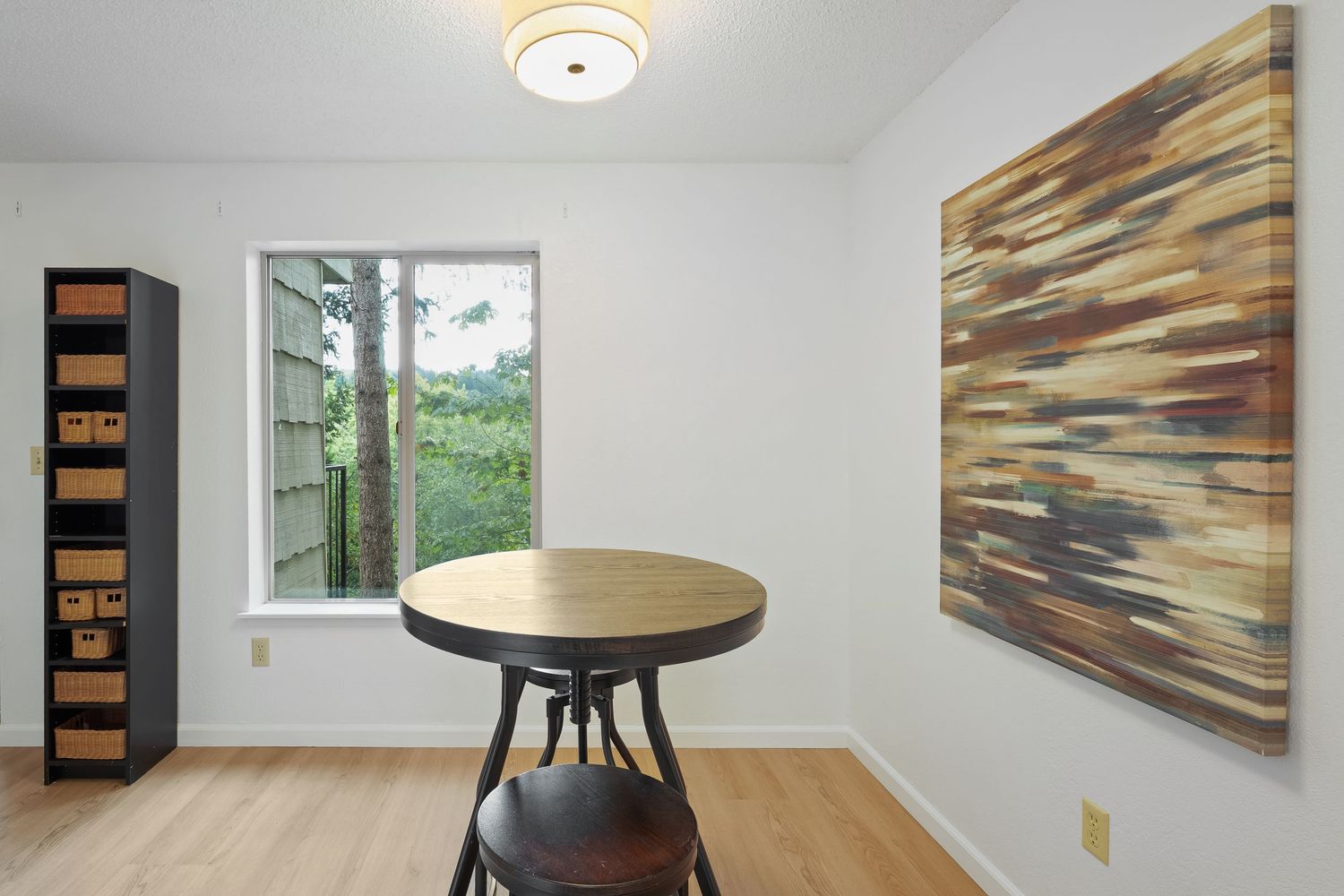 Small dining nook with round wood table, bar stool, abstract wall art, and window overlooking greenery on light hardwood floors.