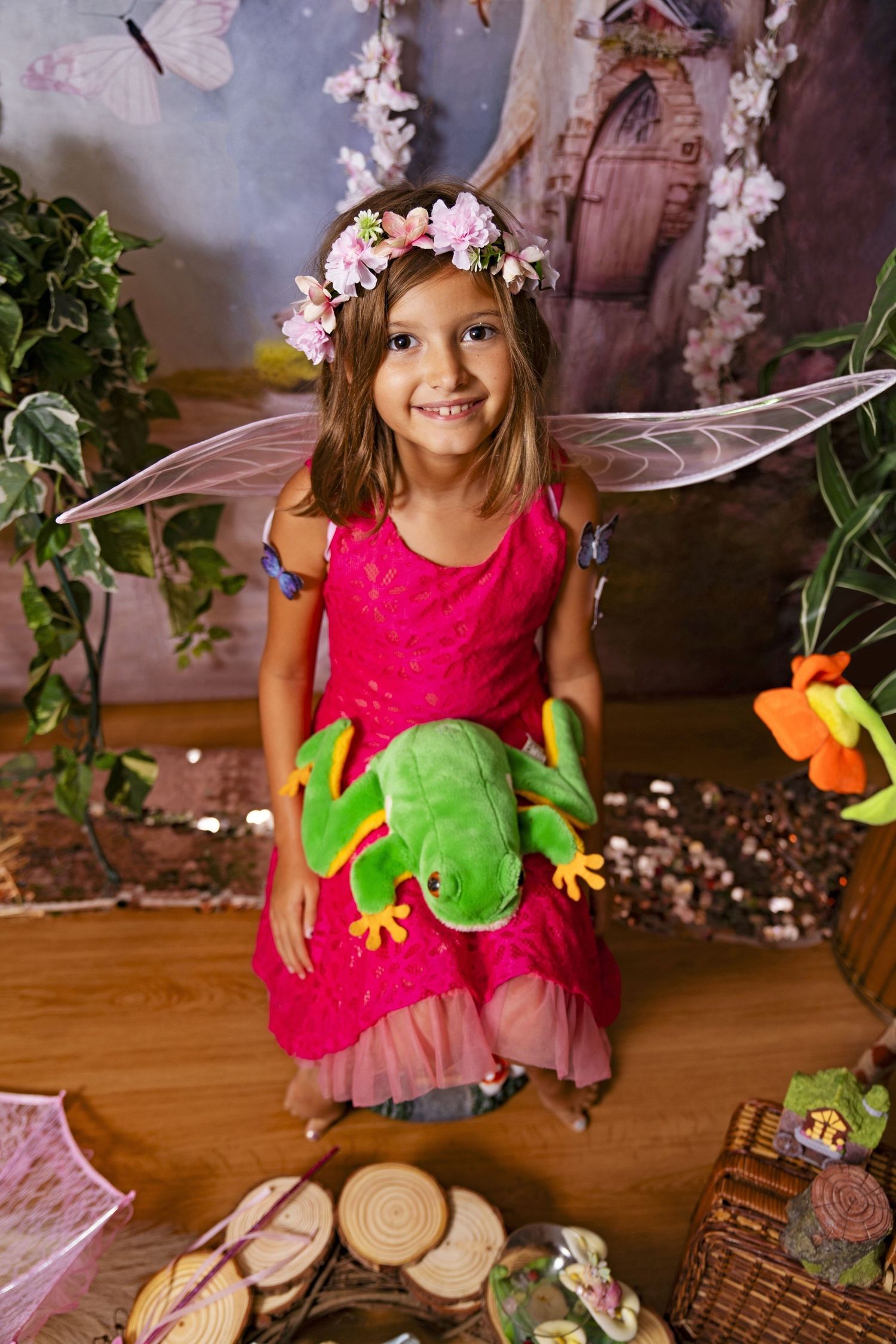 Young girl in pink fairy costume with flower crown holds green frog plush toy in enchanted forest setting.