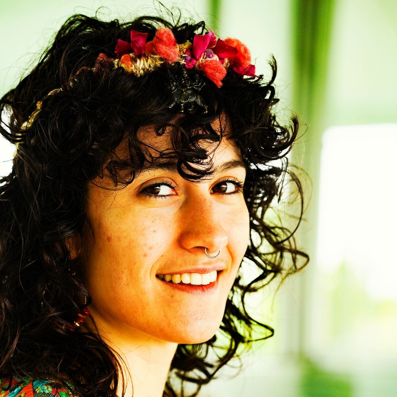 A photo of a girl smiling at the camera, she has black wavy hair and a red flowered flower crown.