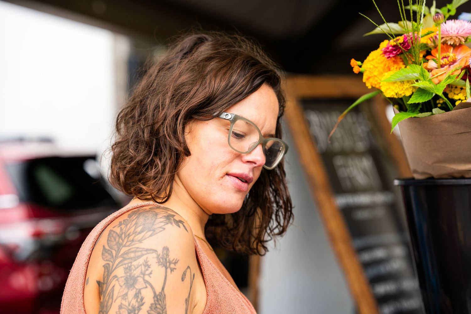 A tattooed woman with curly hair and glasses stands near colorful flowers outdoors.
