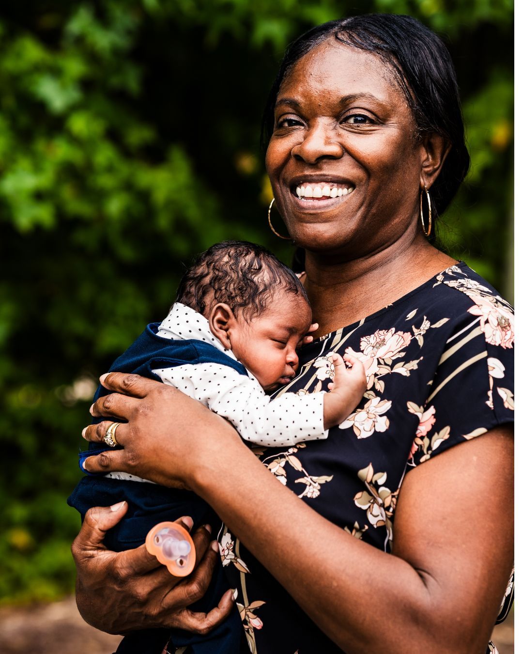 Smiling woman in floral dress holding a sleeping newborn baby outdoors with green foliage background.