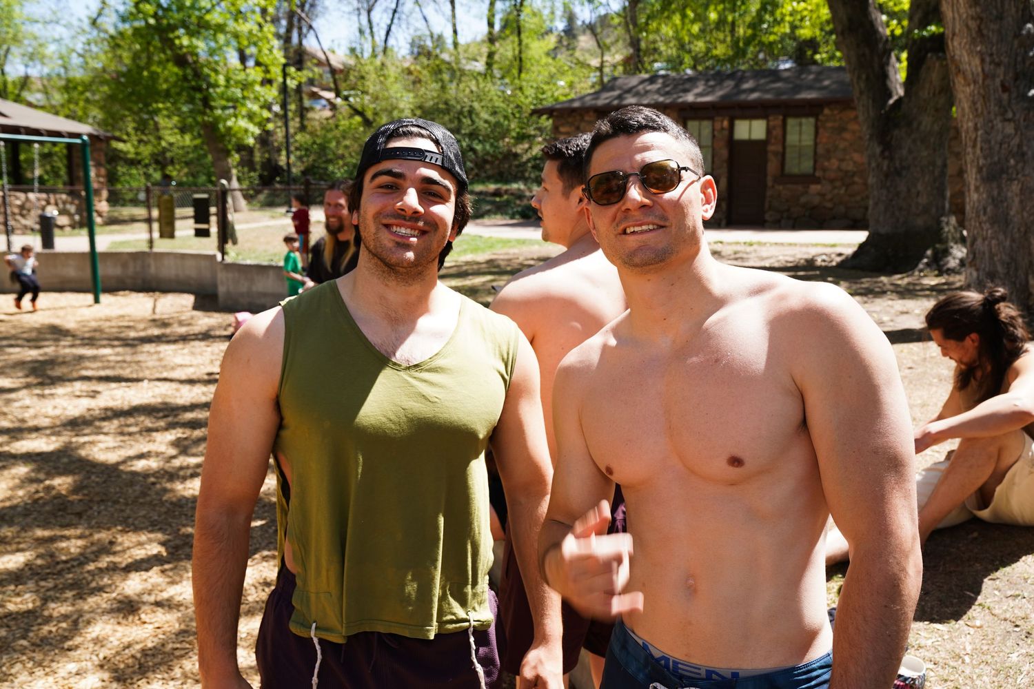 Two shirtless men smiling outdoors in a sunny park setting, one wearing a green tank top and backwards cap.
