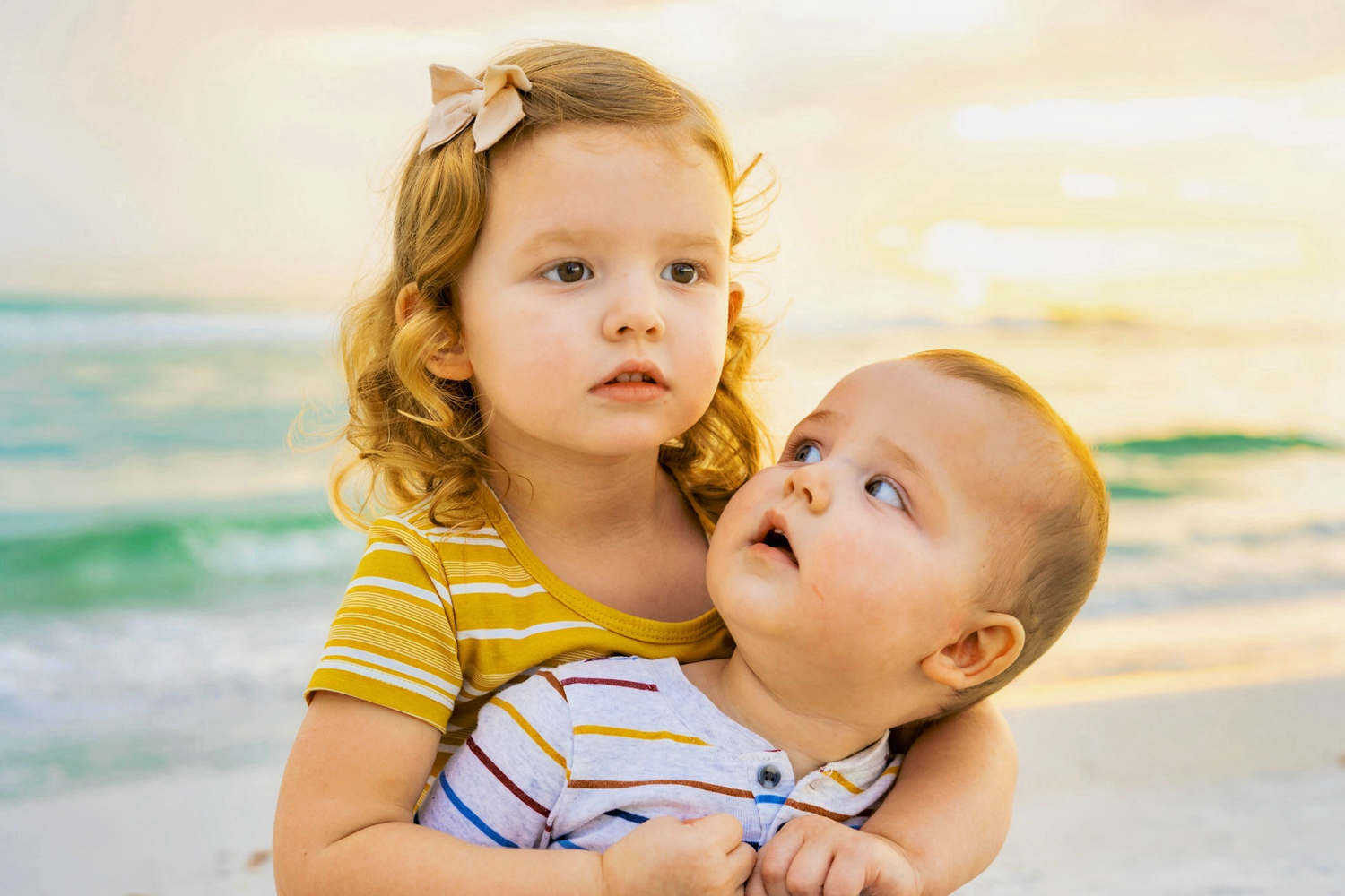 Young girl with curly hair hugs a baby sibling on a sunny beach with turquoise ocean waves in the background.