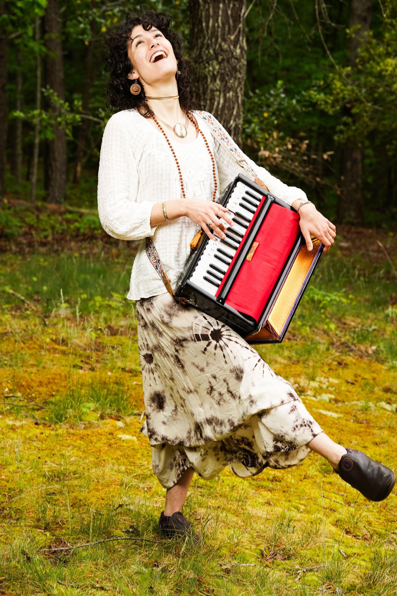 Woman joyfully playing red accordion outdoors in forest wearing bohemian white top and tie-dye skirt.