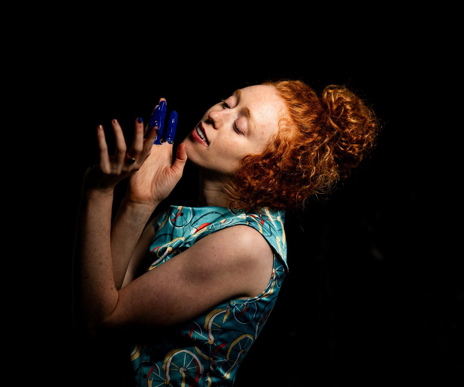 Red-haired woman in teal dress holds blue butterfly near face against dark background.