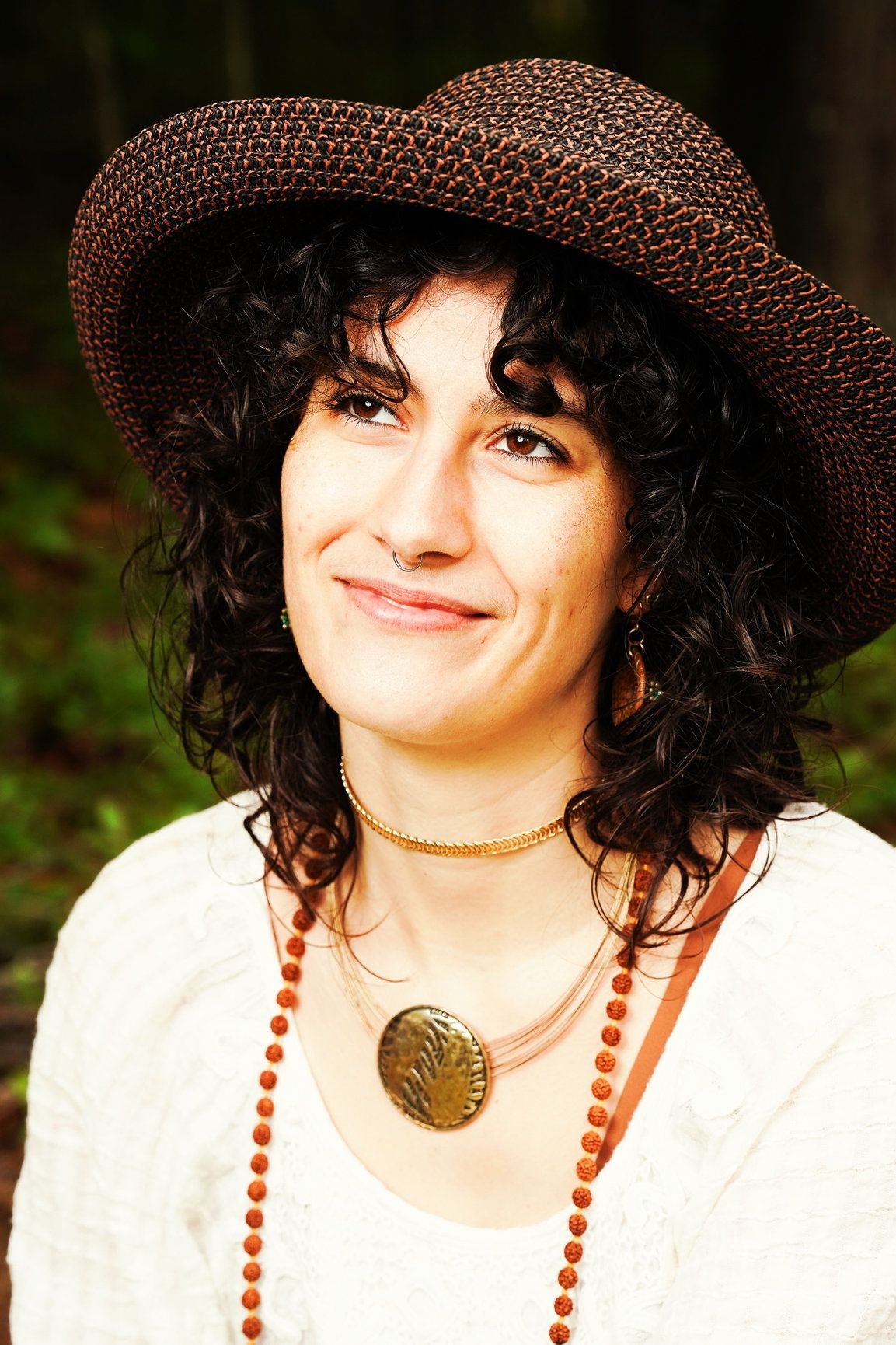 A woman with black curly hair and a brown hat wearing jewelry and a white top.