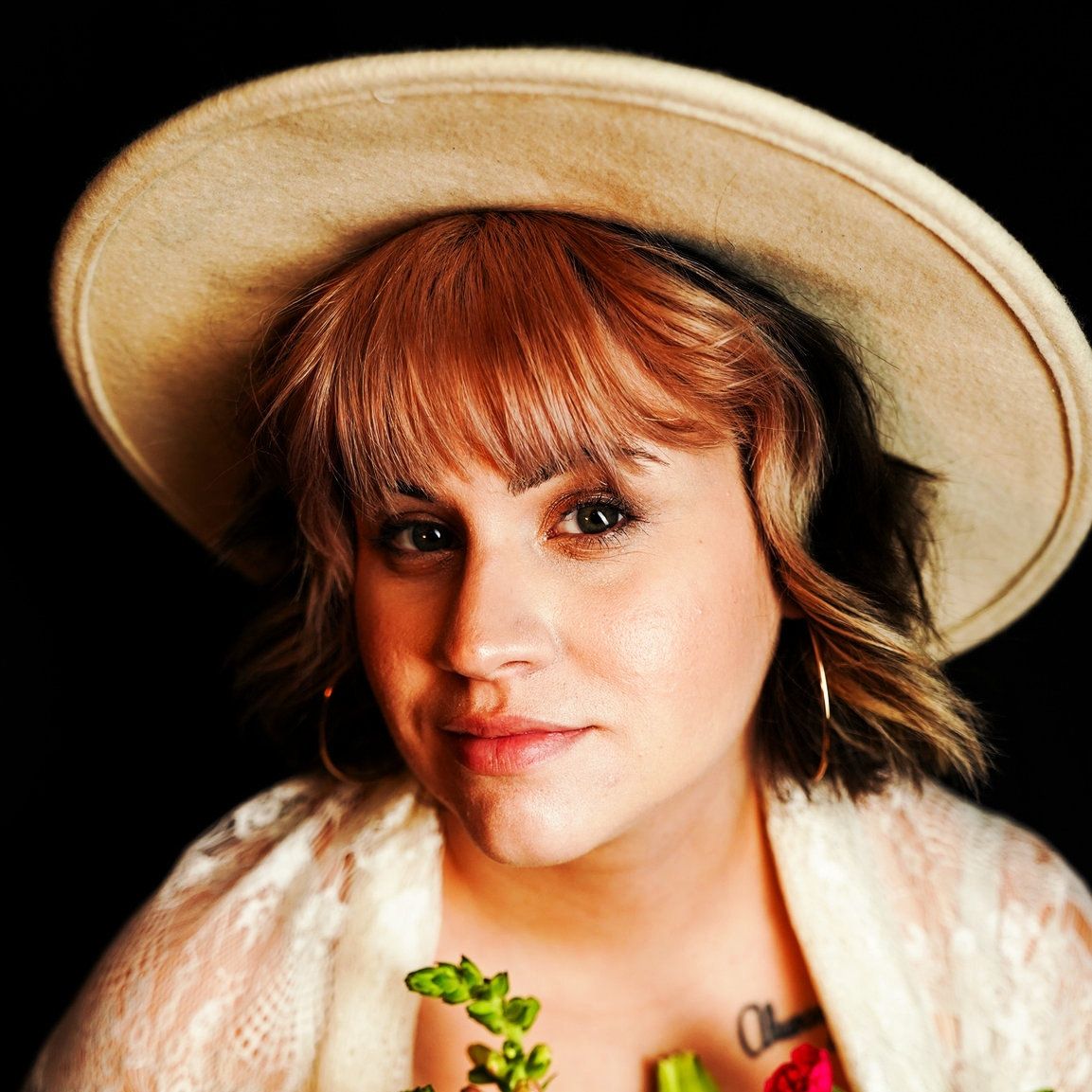 A photo of a woman wearing a tan hat, she has blonde hair and is looking at the camera wearing a white shawl.