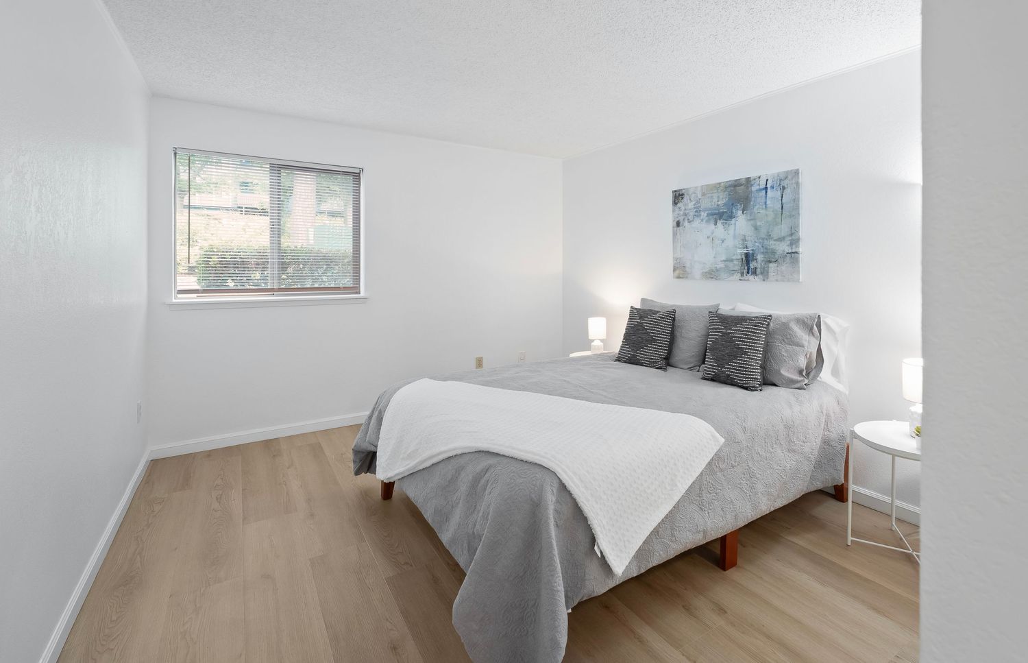 Bright minimalist bedroom with a gray bed, light wood floors, white walls, and a window with natural light.