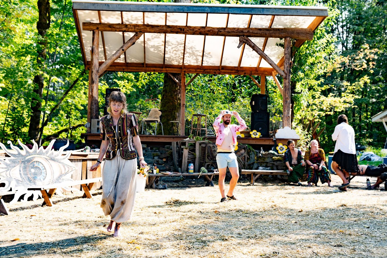 People dancing and socializing outdoors at a rustic woodland festival with a wooden pergola stage.