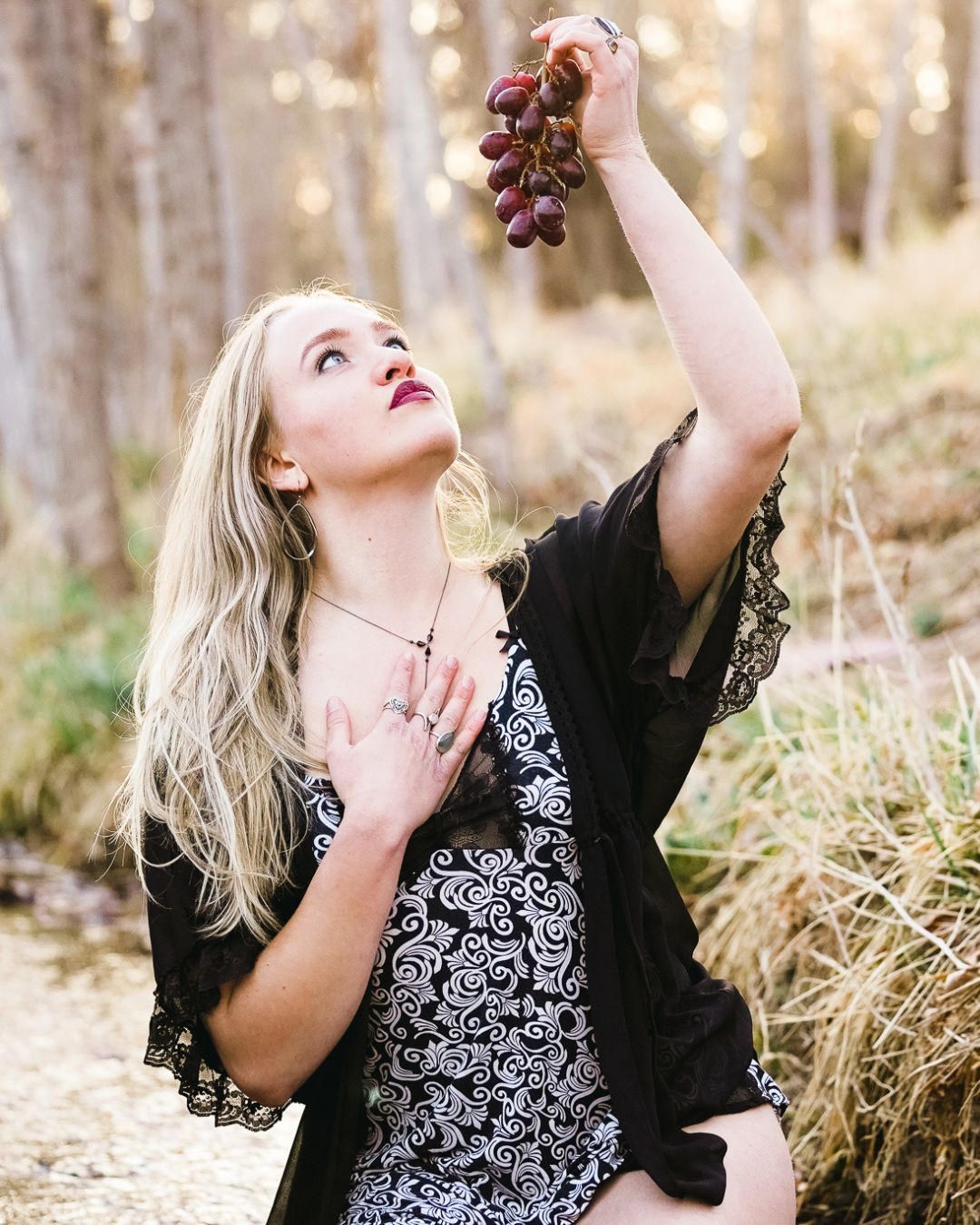 Blonde woman in floral dress and black lace kimono holds red grapes overhead in a sunlit forest setting.