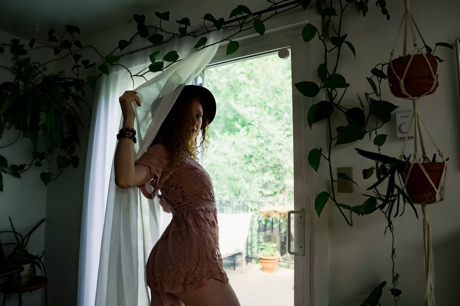 Woman in pink dress pulling back white curtain by open door surrounded by hanging green plants indoors.