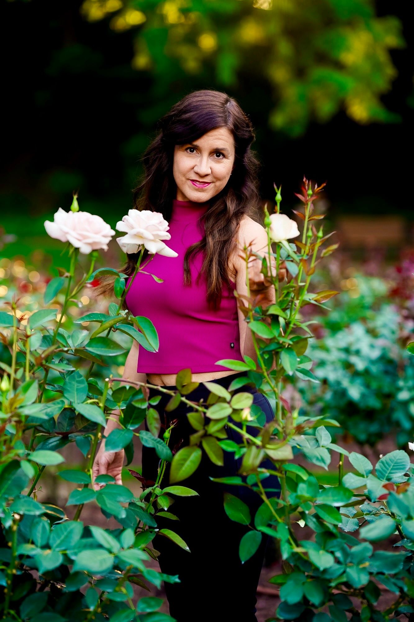 Person in magenta turtleneck poses among blooming white roses in a lush garden setting.