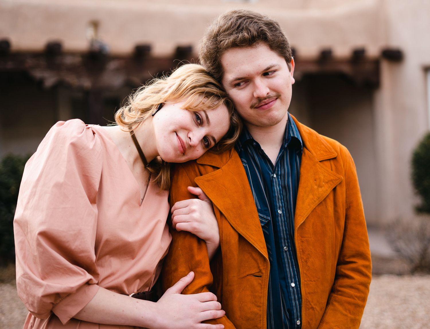 A couple in orange and pink coats share tender moments while posing together outdoors.
