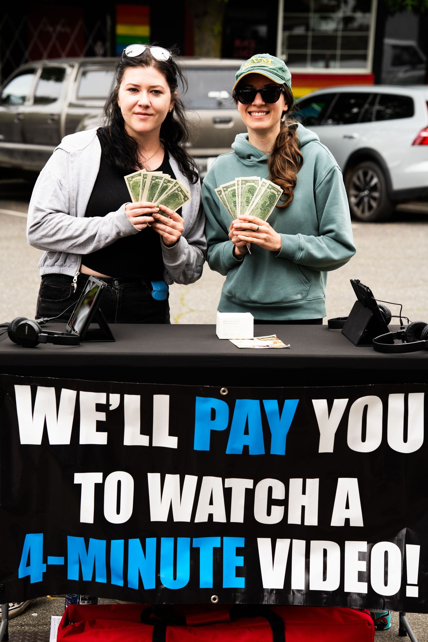 Two women holding cash at a table with a sign reading We'll Pay You to Watch a 4-Minute Video.