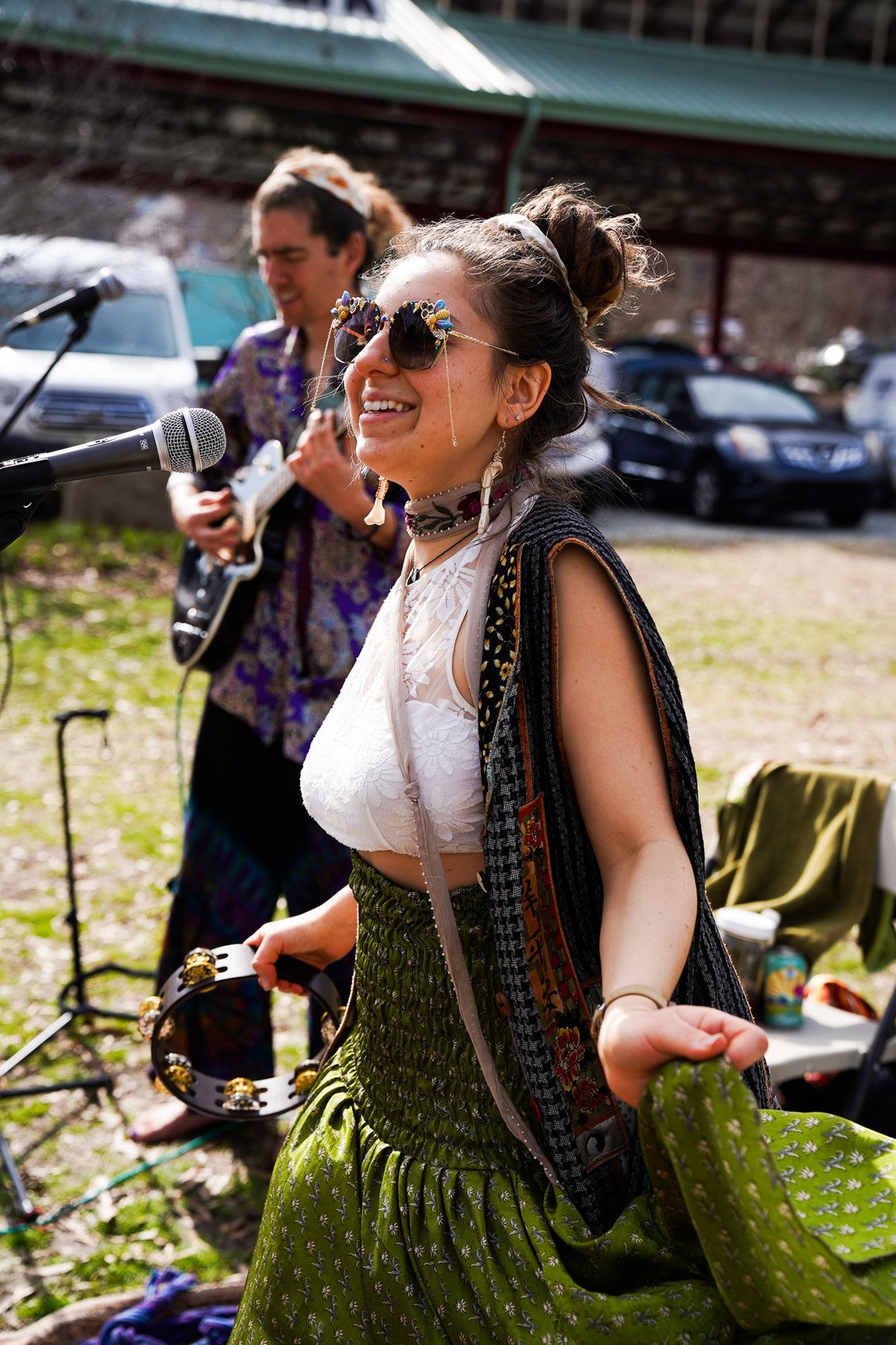 A woman wearing a colorful vest, bright glasses, and earrings dances and sings into a microphone.