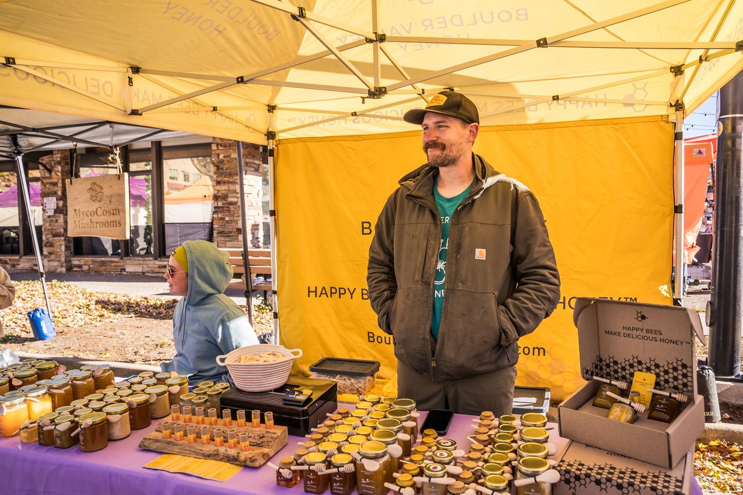 Happy Bees vendor smiling at his honey booth at Boulder Farmers Market, featuring local Colorado honey.