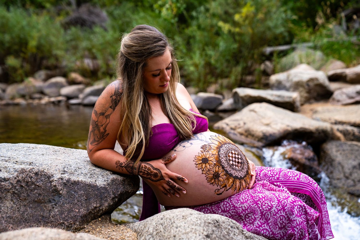 A pregnant woman in purple attire sits on rocks by a stream with henna art adorning her belly.