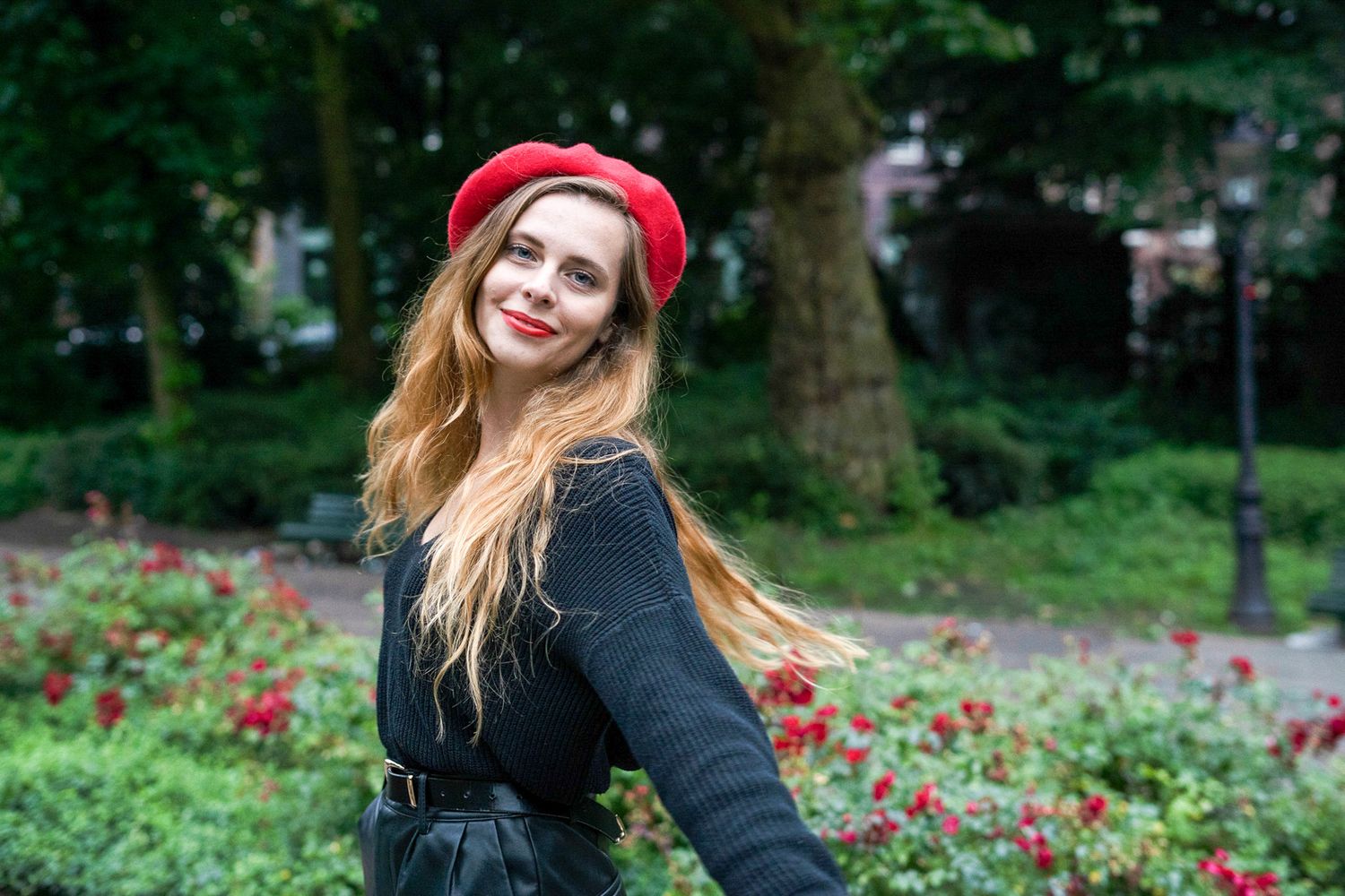 A photo of a girl wearing black and a bright red hat with bright red lipstick and roses behind her.